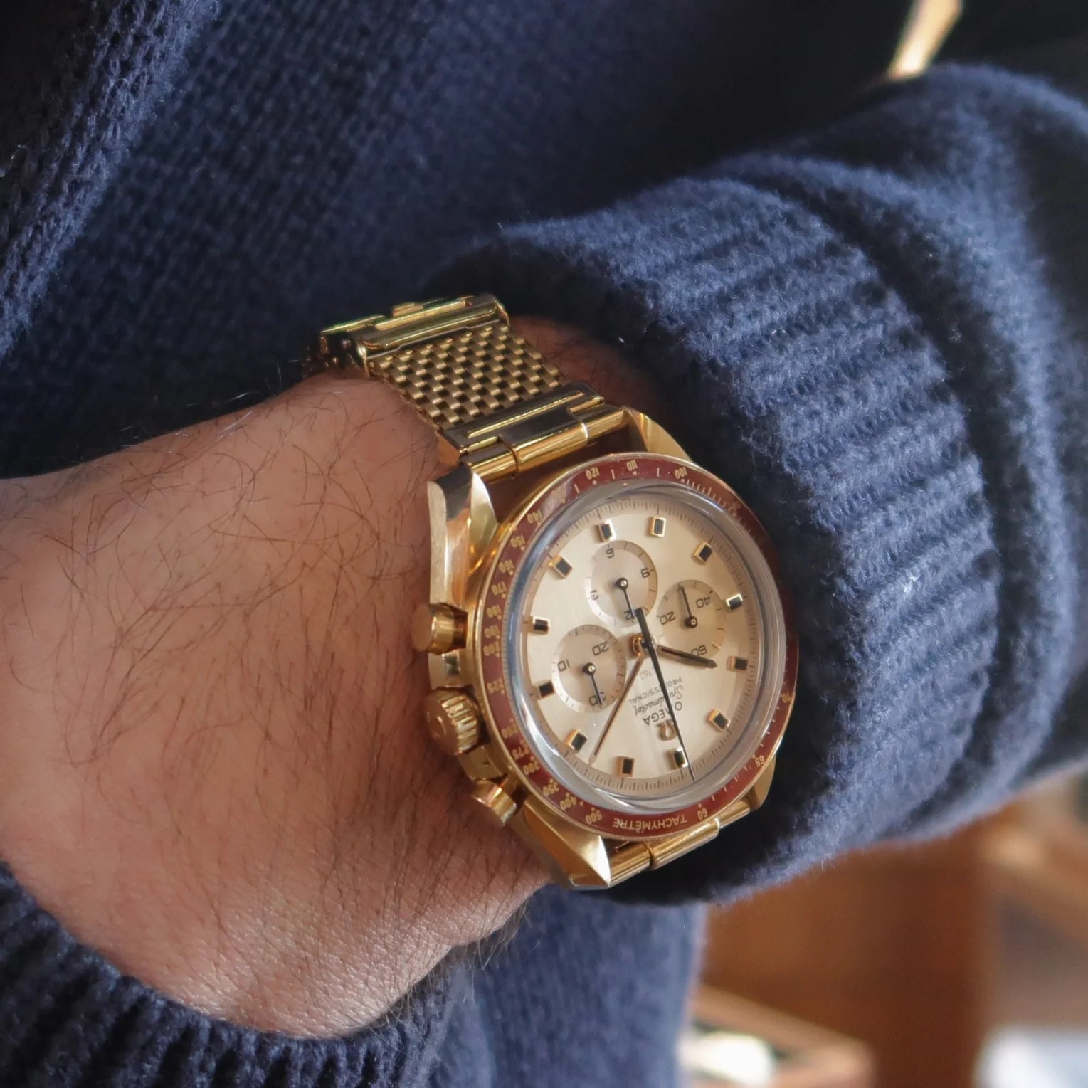 Gold plated chronograph watch with mesh bracelet worn on wrist over blue sweater