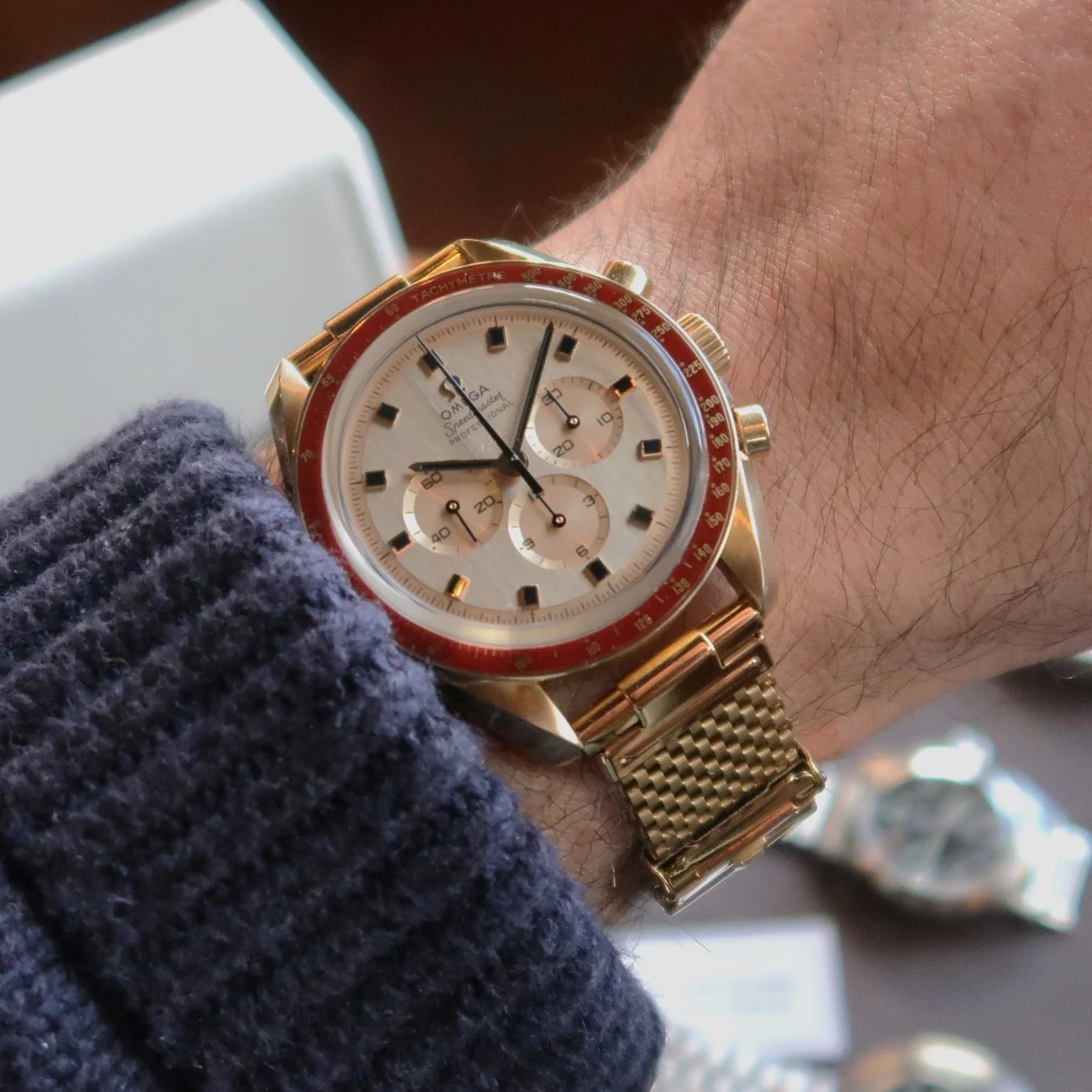 Gold plated chronograph watch with cream dial, red bezel, and mesh bracelet worn on wrist with blue sweater