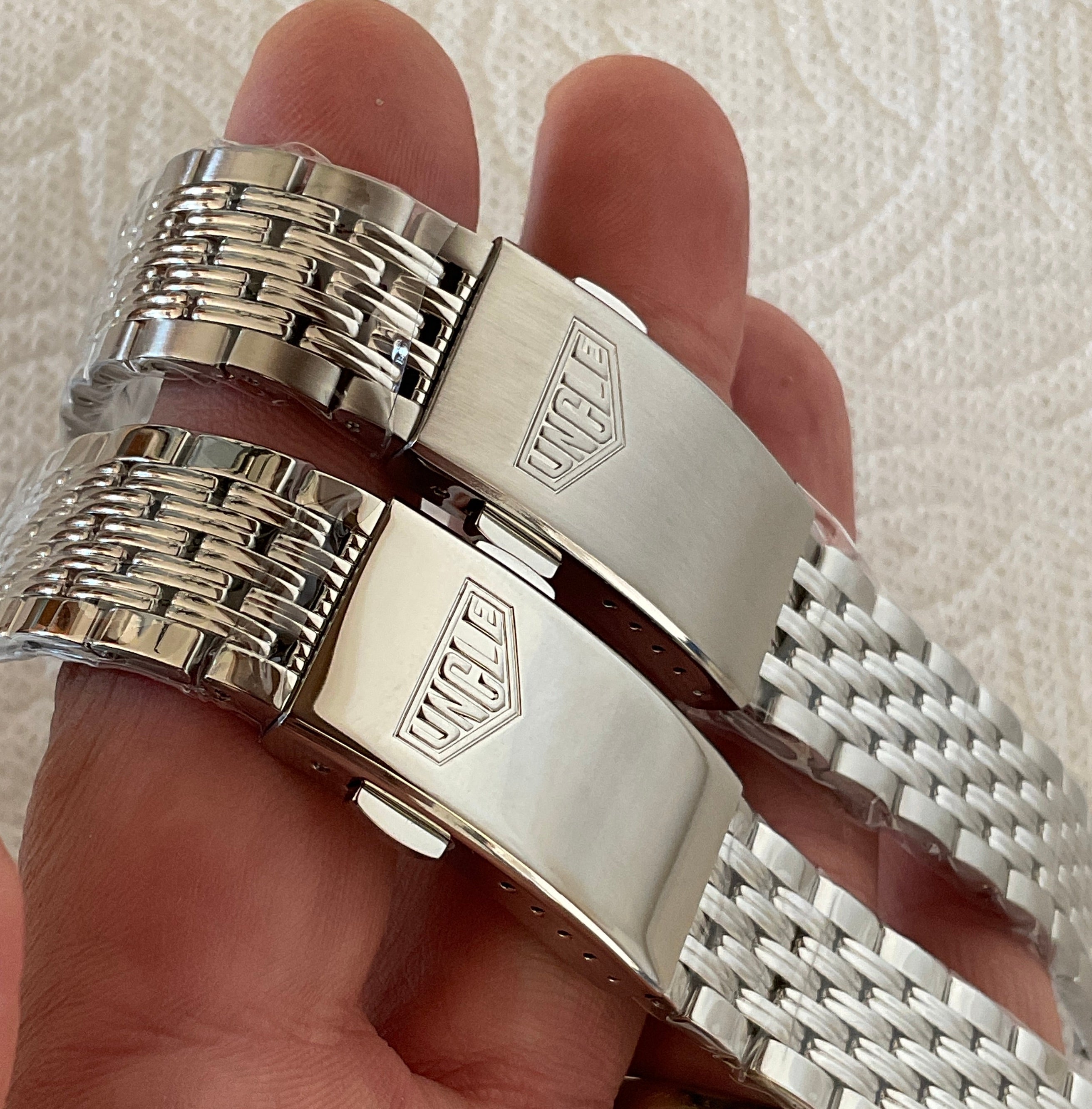 Pair of stainless steel bracelets with polished links and branded buckle clasps, displayed on hand against textured background