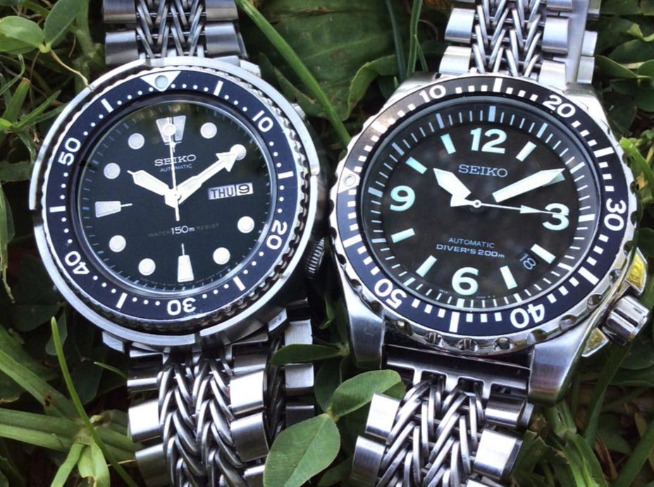Two Seiko automatic dive watches with steel bracelets displaying black dials and luminous hour markers on green foliage background