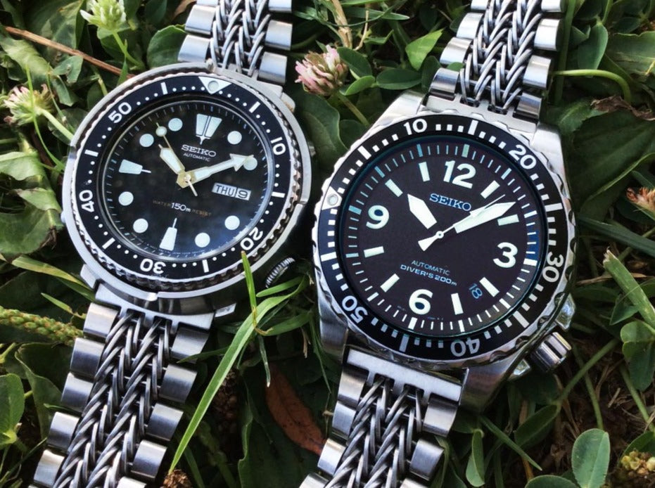 Two Seiko automatic dive watches with stainless steel Razor-Wire bracelets on green foliage background