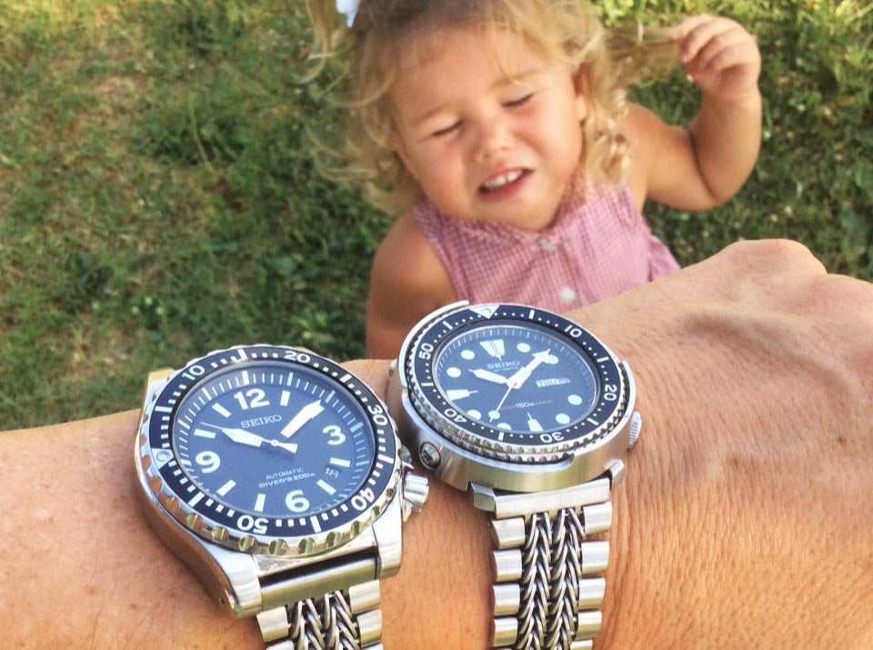 Two stainless steel dive watches with metal bracelets worn on wrist, child in pink shirt playing in background with green hedges