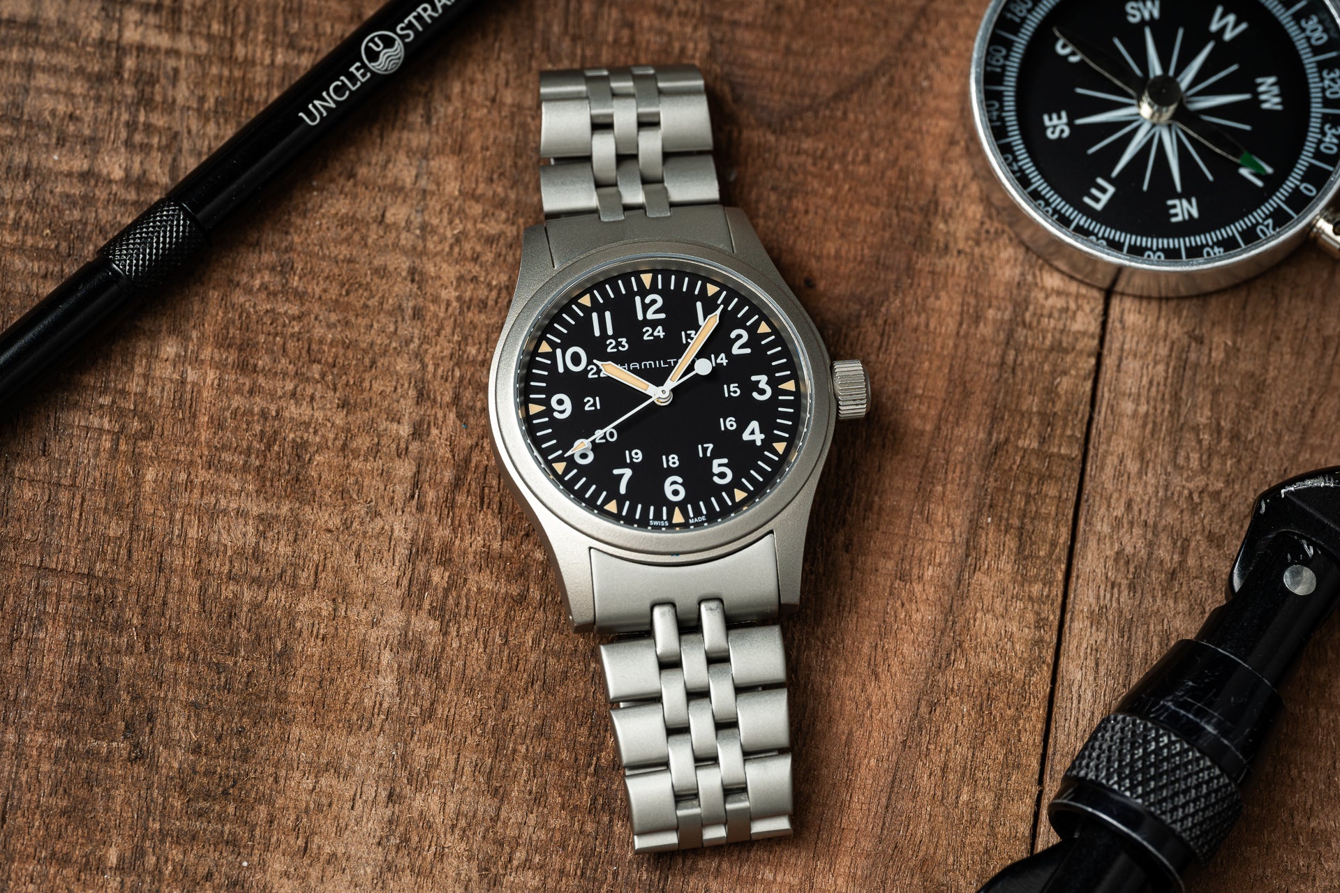 Hamilton Khaki mechanical watch with black dial and 24-hour markers, stainless steel bracelet on wooden surface with compass and tactical gear