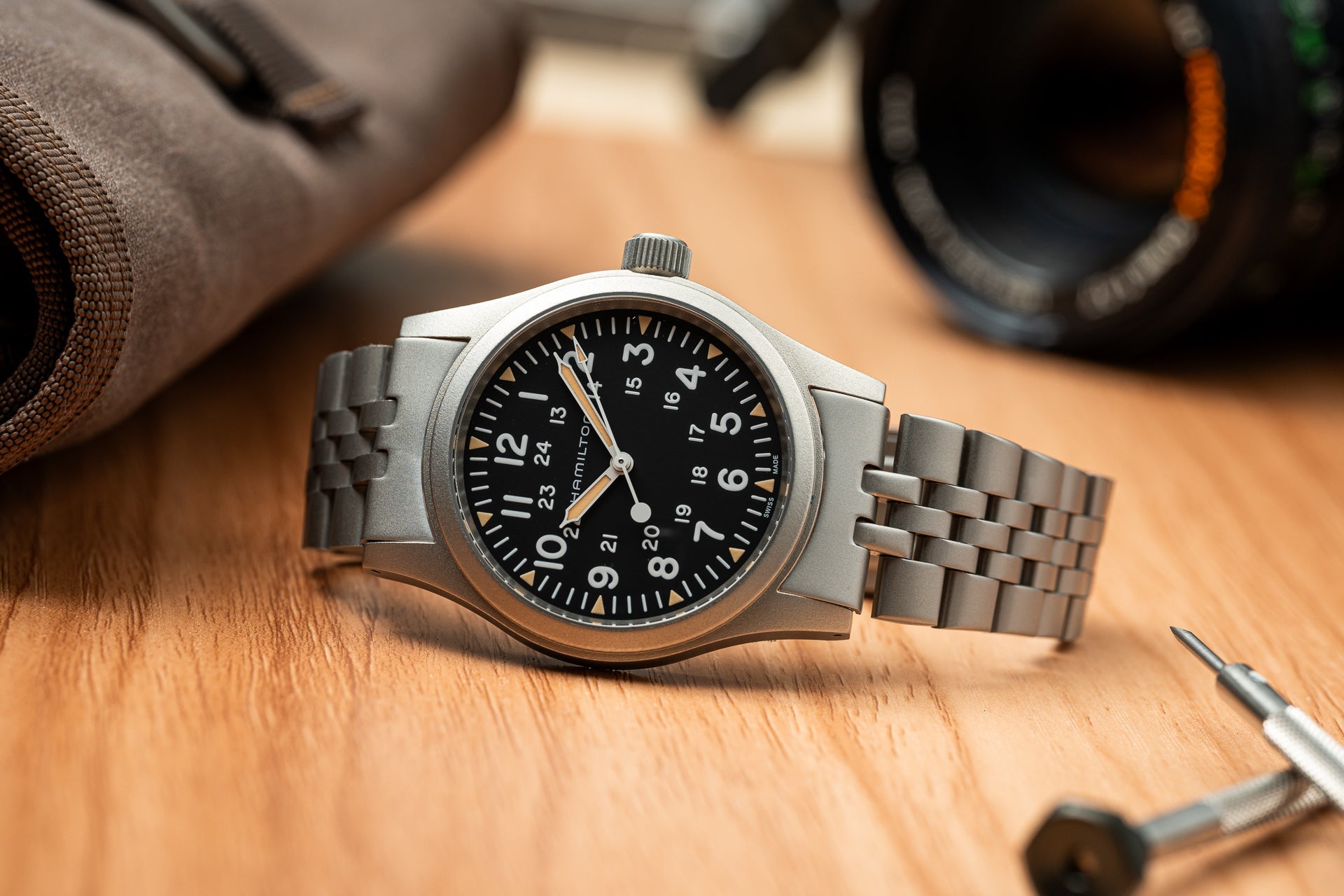 Stainless steel mechanical watch with black dial, Arabic numerals, and metal bracelet on wooden surface with camera gear
