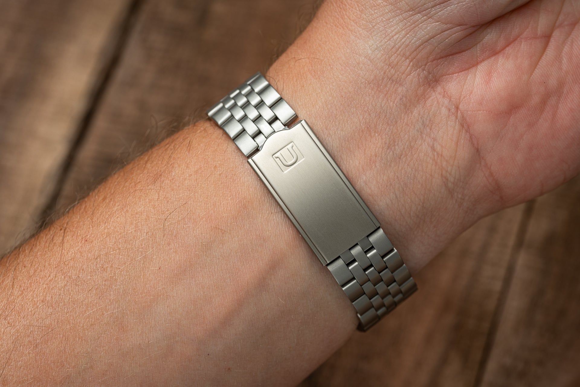Stainless steel metal bracelet with rectangular logo plaque worn on wrist against brown fabric background