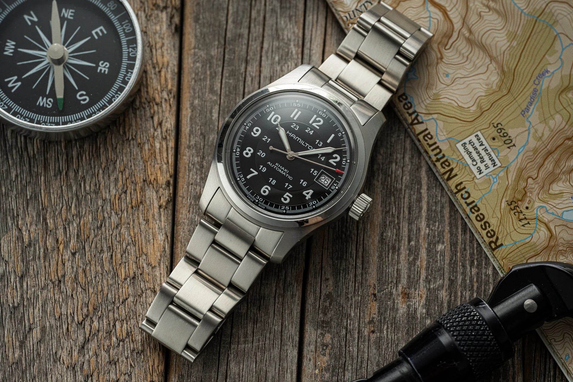 Hamilton Khaki Field automatic stainless steel watch with black dial, date window, and metal bracelet on wooden surface with compass and topographic map