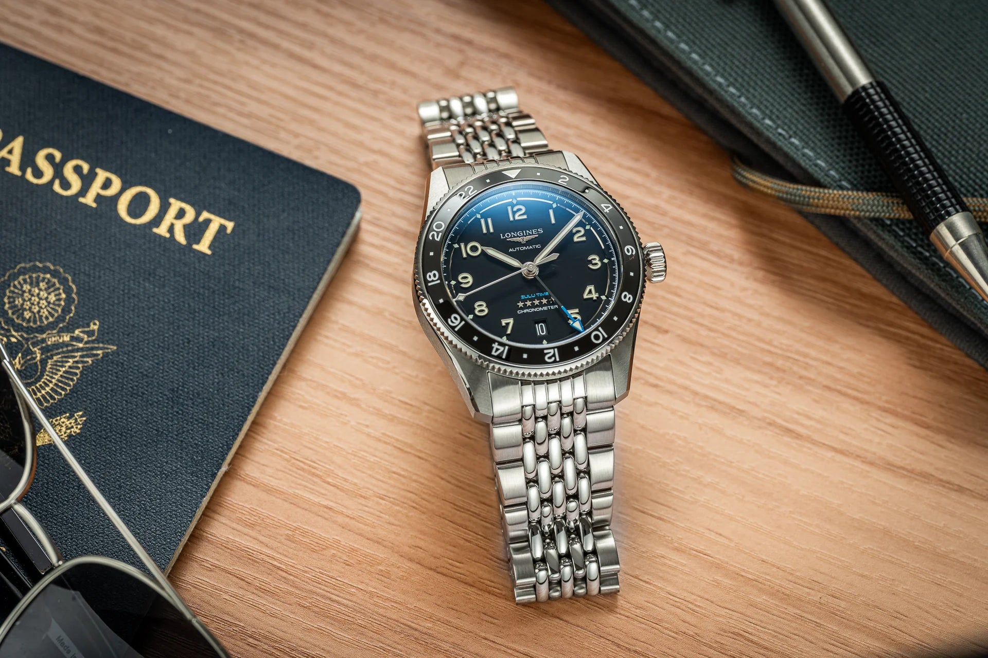 Longines Spirit Zulu Time 39mm automatic watch with black dial, steel bracelet, and rotating GMT bezel on wooden desk with passport