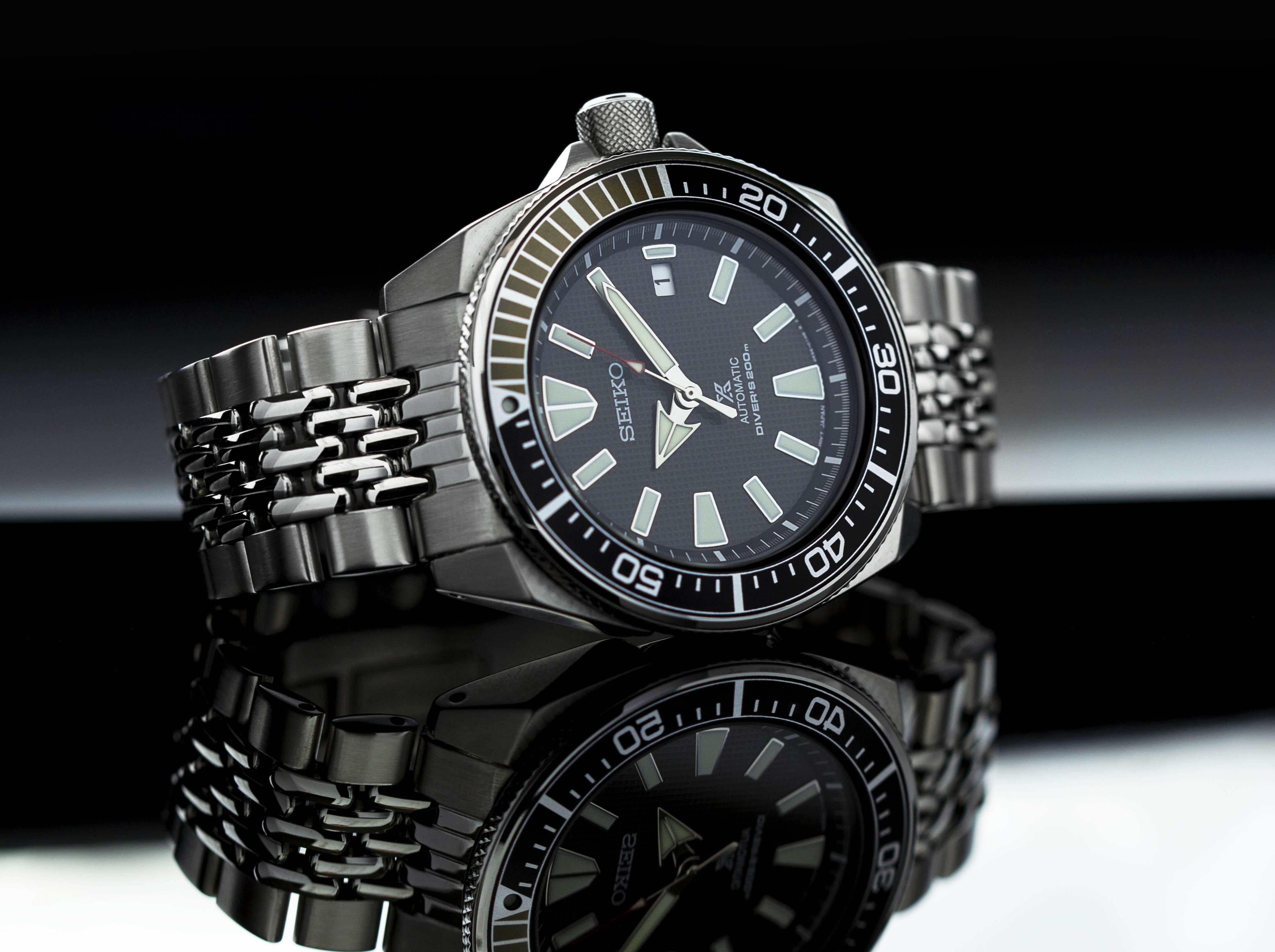 Seiko Samurai automatic dive watch with black dial, luminous indices, stainless steel beads of rice bracelet, and rotating bezel on reflective surface