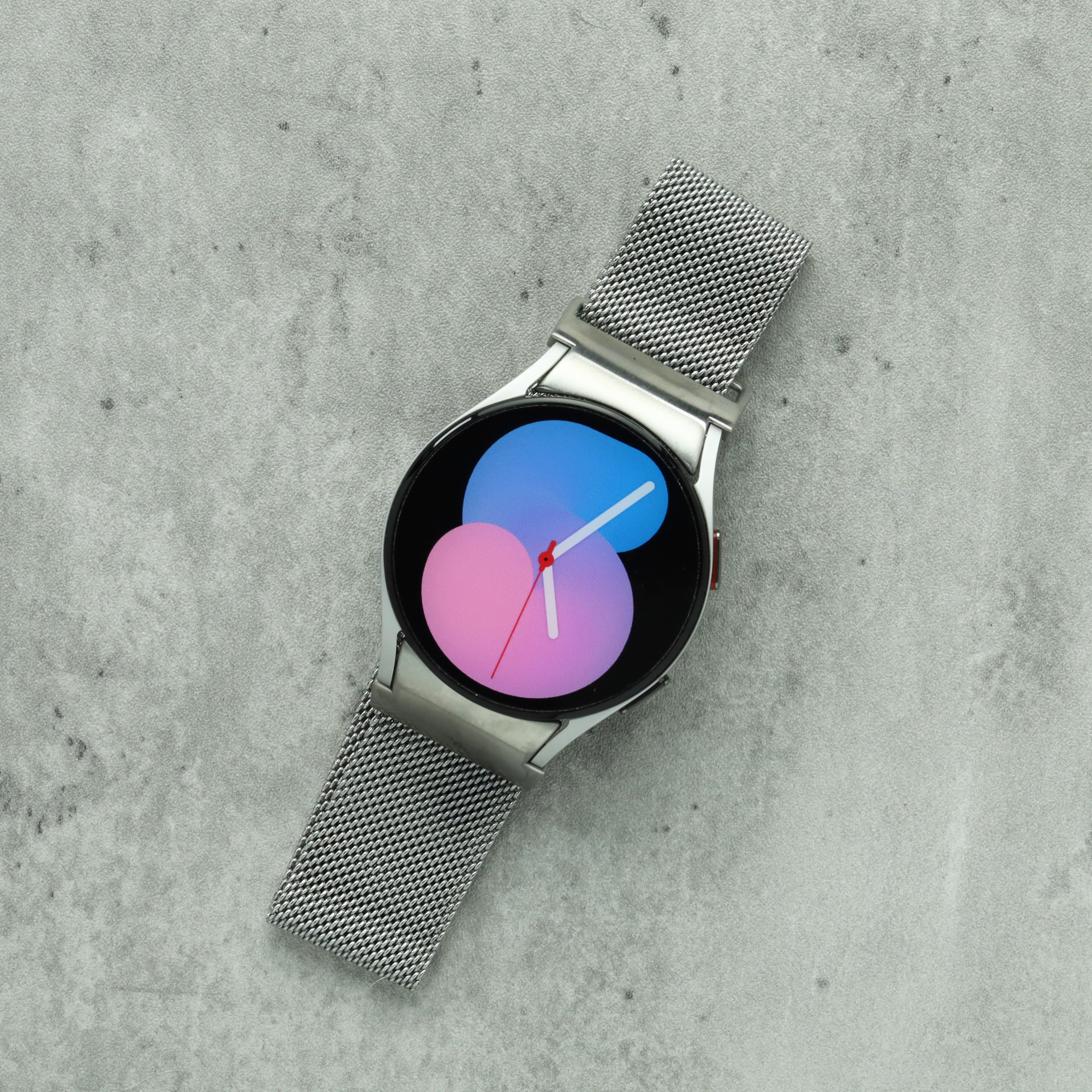 Silver Milanese mesh smartwatch with gradient blue-pink display and analog watch face on concrete background