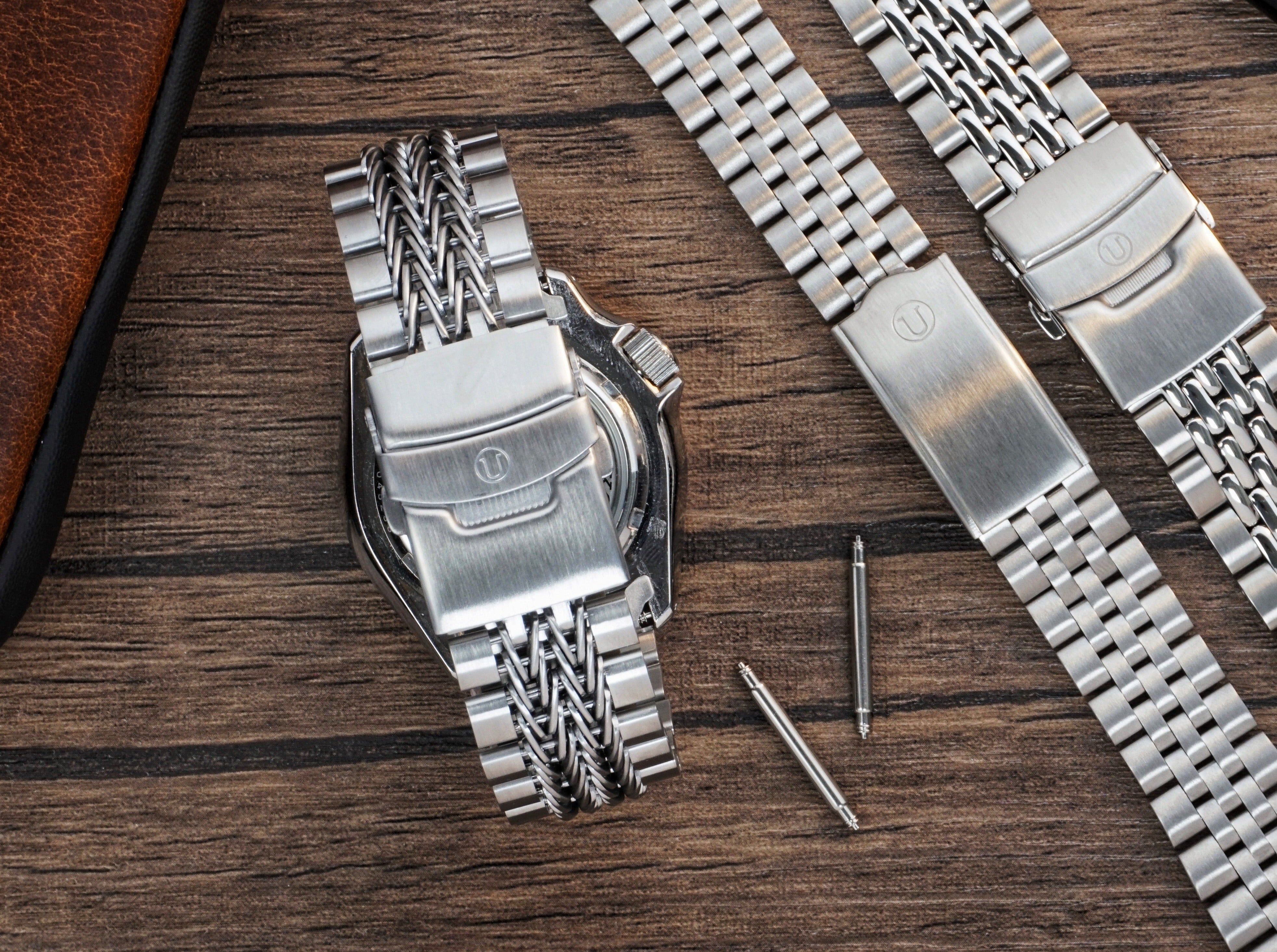 Stainless steel watch bracelets with twisted mesh and jubilee links displayed on wooden surface with watch repair tools