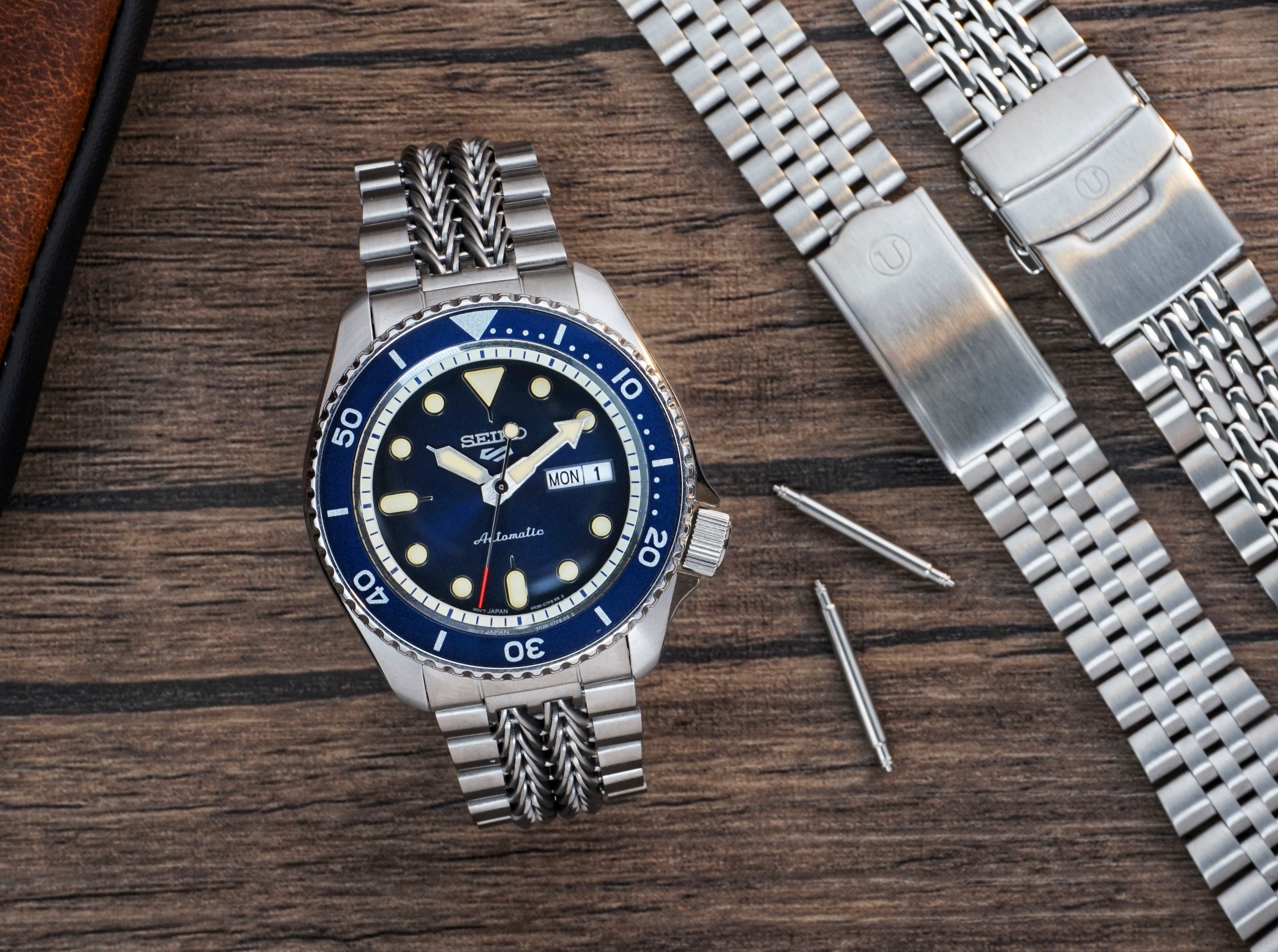 Seiko automatic dive watch with blue dial and bezel, stainless steel bracelet, day-date window, displayed with extra watch straps on wooden surface