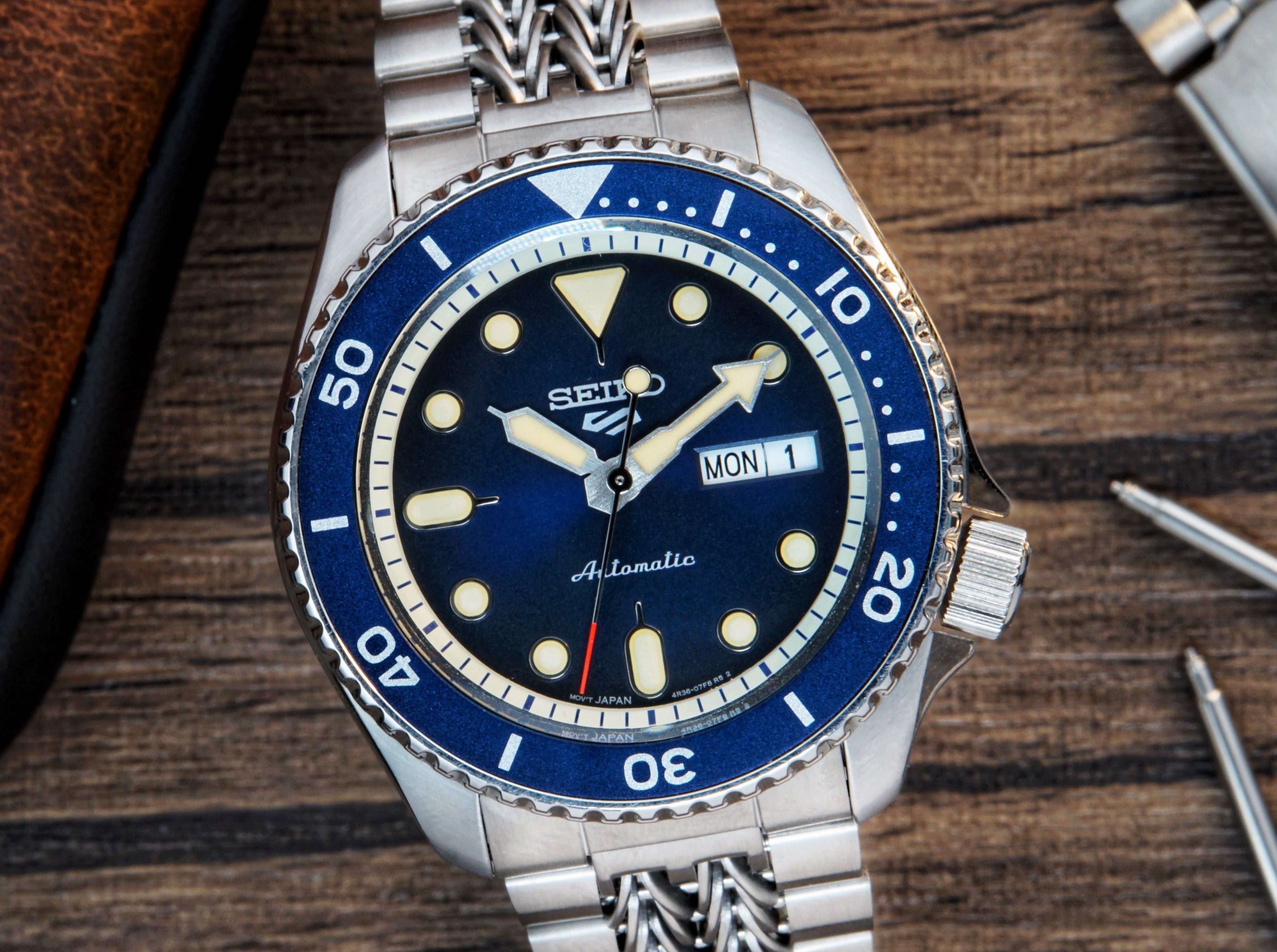 Seiko 5 Sports automatic dive watch with blue dial, stainless steel bracelet, day-date window, and luminous hands on wooden background