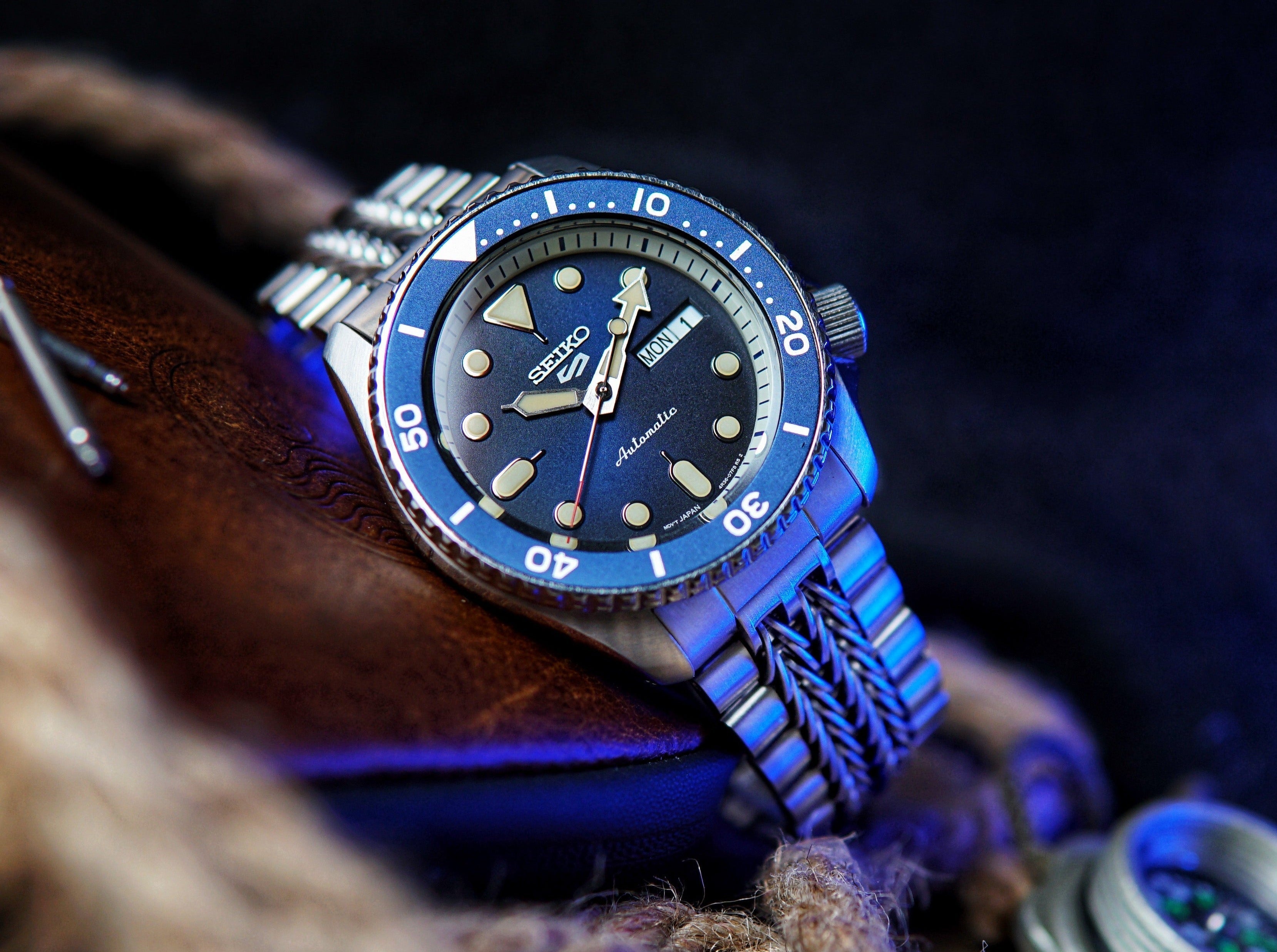 Seiko 5 Sports automatic watch with blue dial, rotating bezel, day-date window, and metal bracelet on dark background