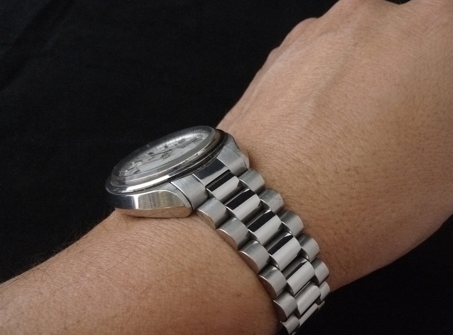 Men's stainless steel watch with oval face and metal bracelet worn on wrist against black background
