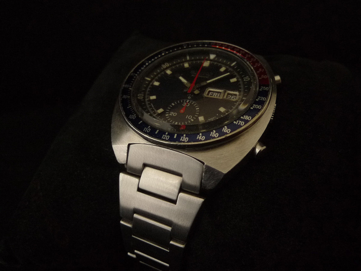 Vintage Seiko 6139 chronograph watch with black dial, red hands, stainless steel tapered H-link bracelet, day-date window, and tachymeter bezel