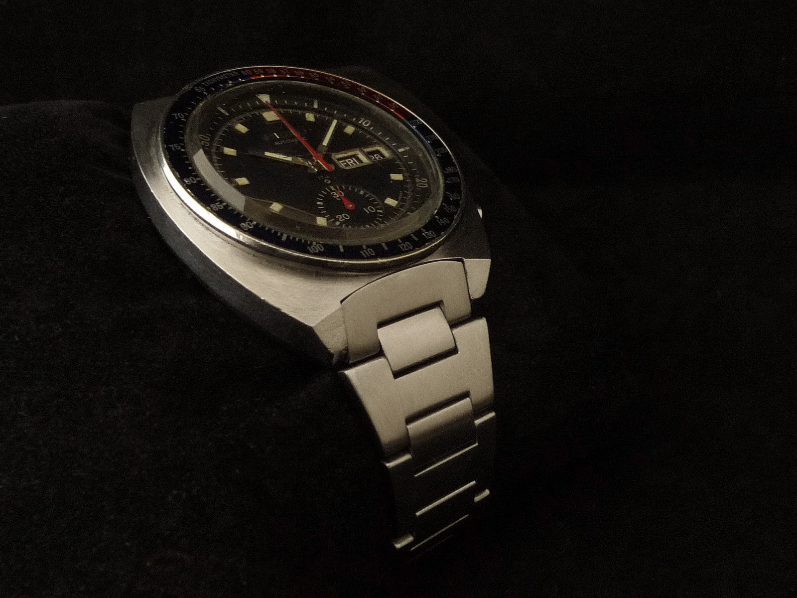 Vintage Seiko 6139 chronograph watch with black dial, cream indices, red seconds hand, tapered H-link metal bracelet on dark background