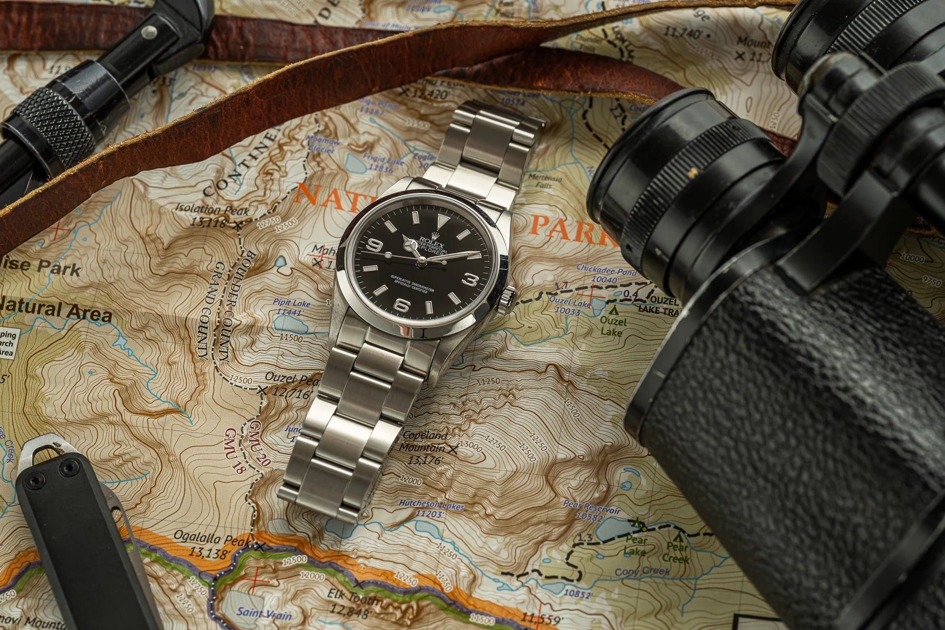 Silver stainless steel Rolex Explorer watch with black dial on topographic map surrounded by binoculars and leather camera strap