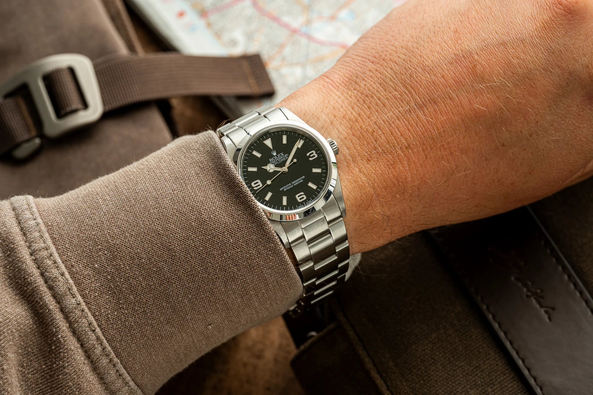 Stainless steel sports watch with black dial worn on wrist over beige fabric, displayed with travel map and brown bag in background