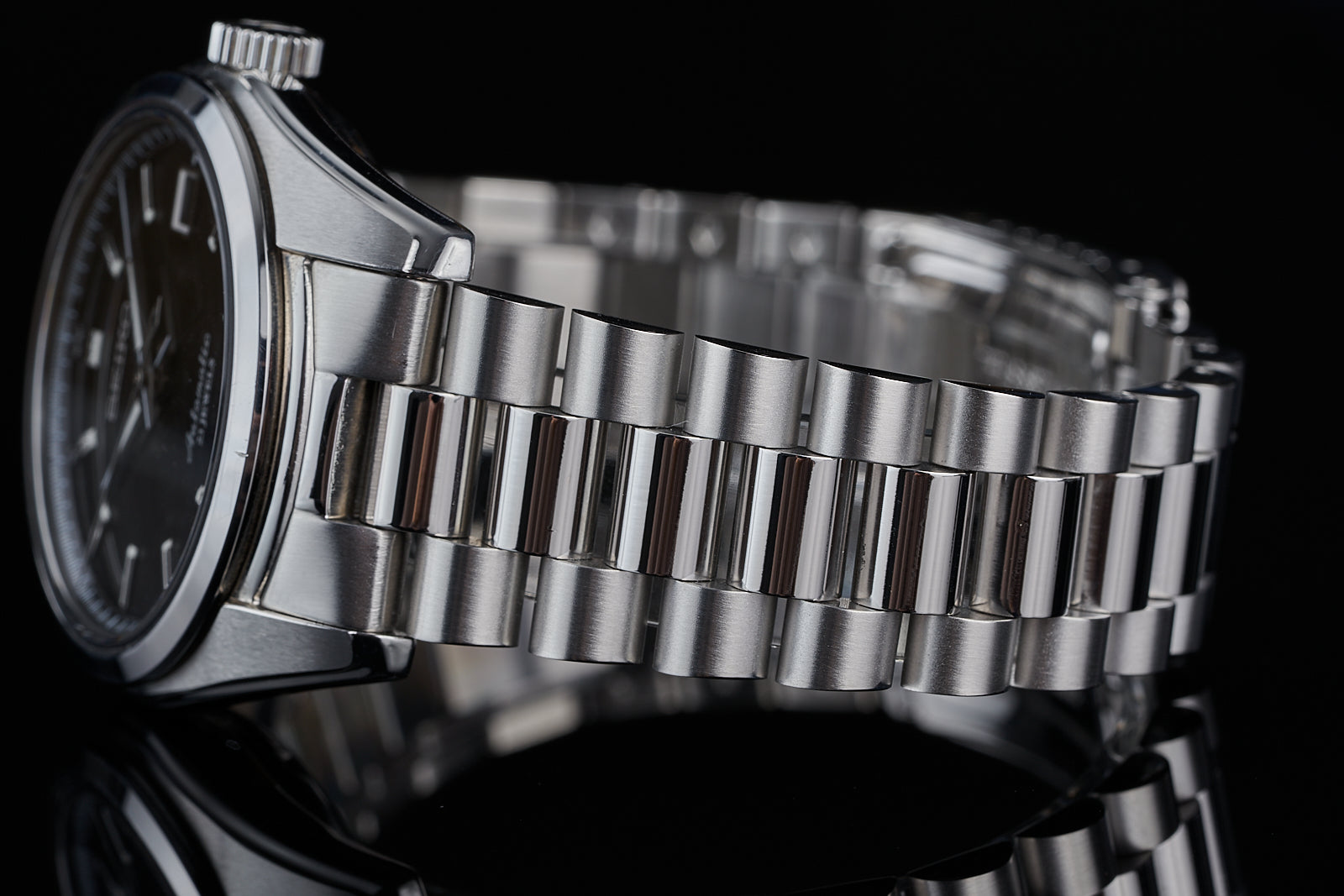 Stainless steel three-link bracelet watch with black dial displayed on reflective surface against black background