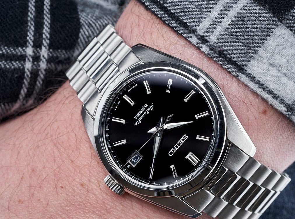 Men's Seiko SARB automatic watch with black dial and stainless steel bracelet worn on wrist with plaid sweater