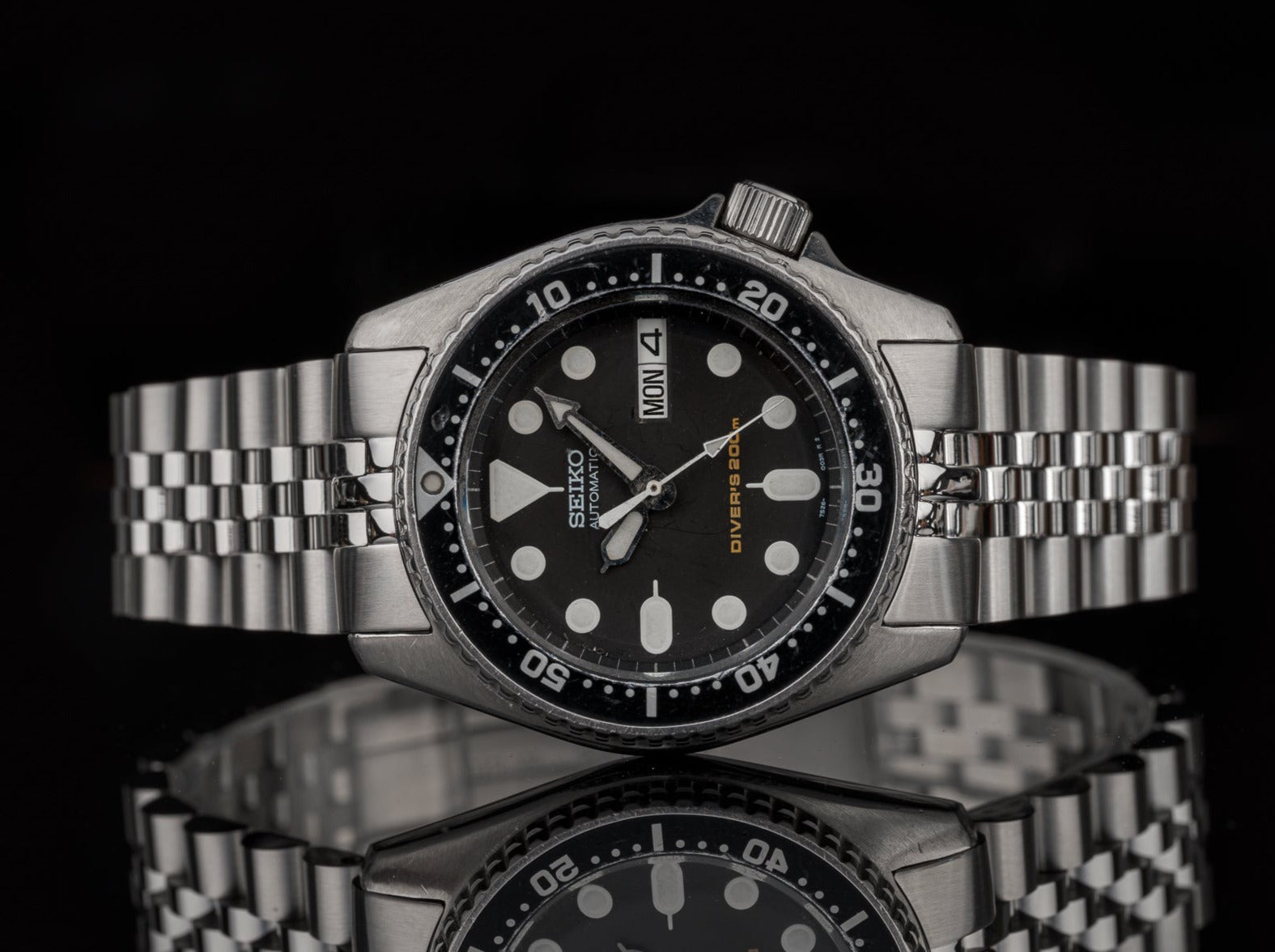 Seiko automatic dive watch with black dial, stainless steel bracelet, day-date window, and rotating bezel on reflective surface