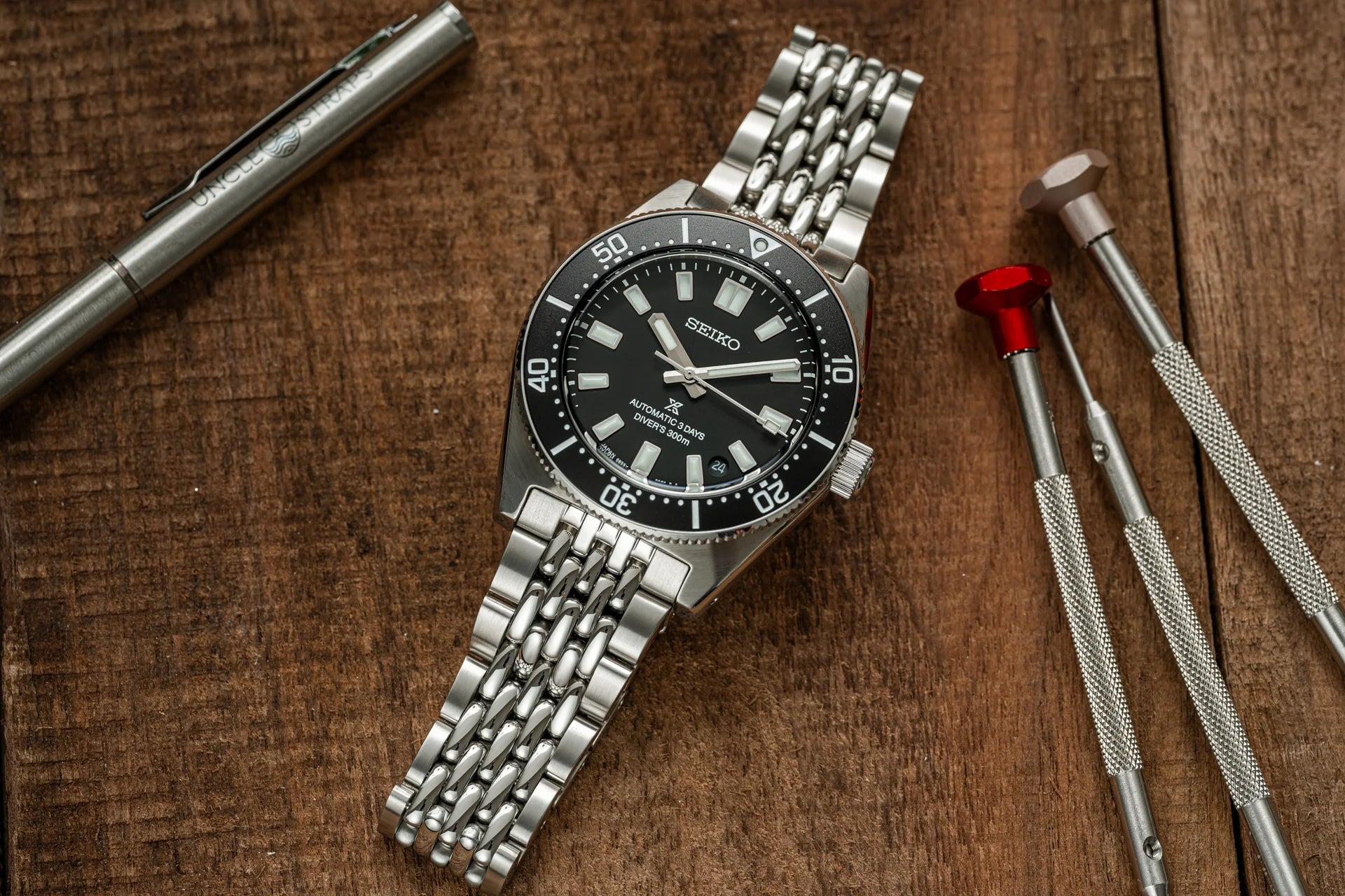 Seiko automatic dive watch with black dial and stainless steel beads of rice bracelet, displayed with watch tools on wooden background