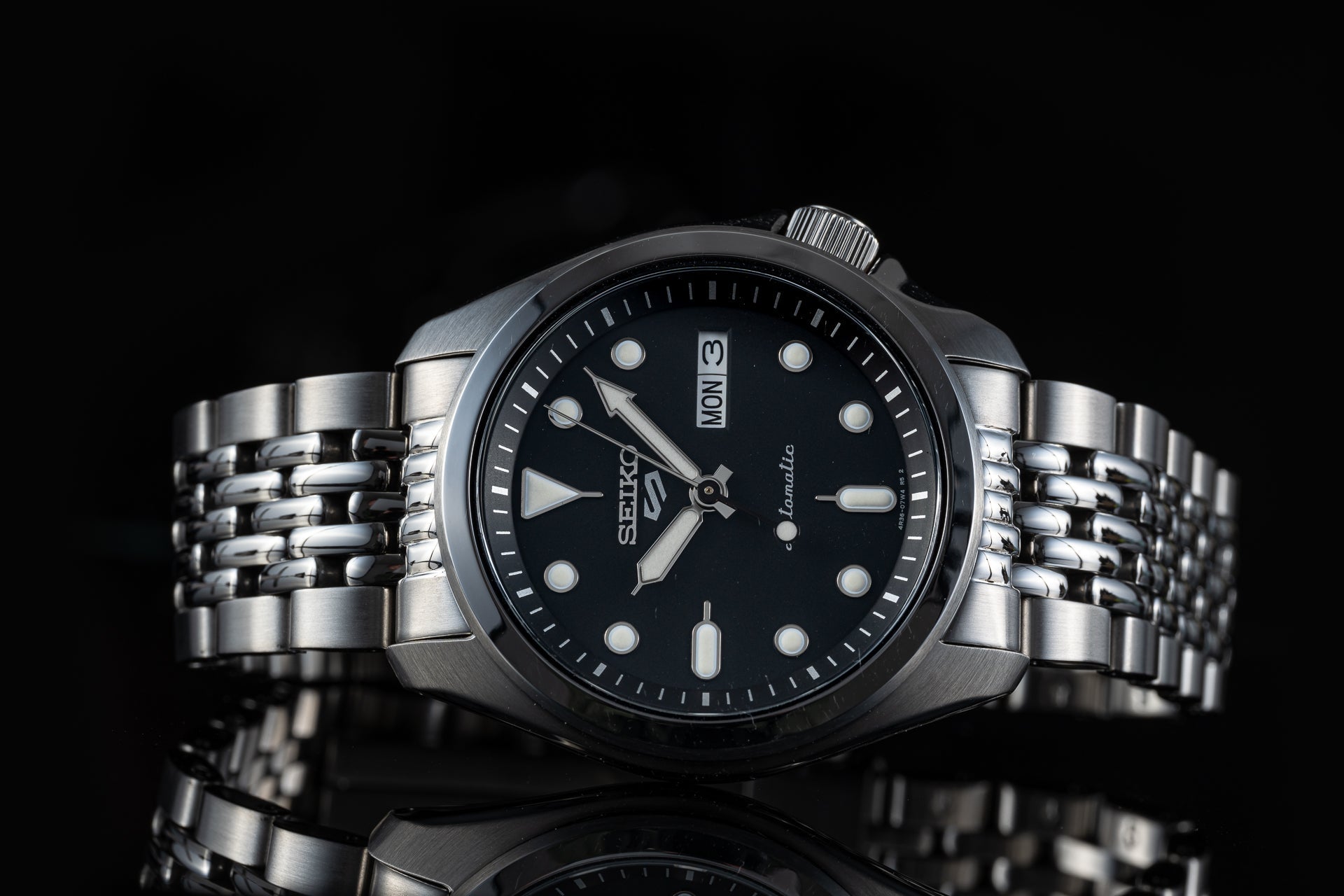 Seiko 5 Sports automatic watch with black dial, day-date window, luminous markers, stainless steel beads of rice bracelet on black background