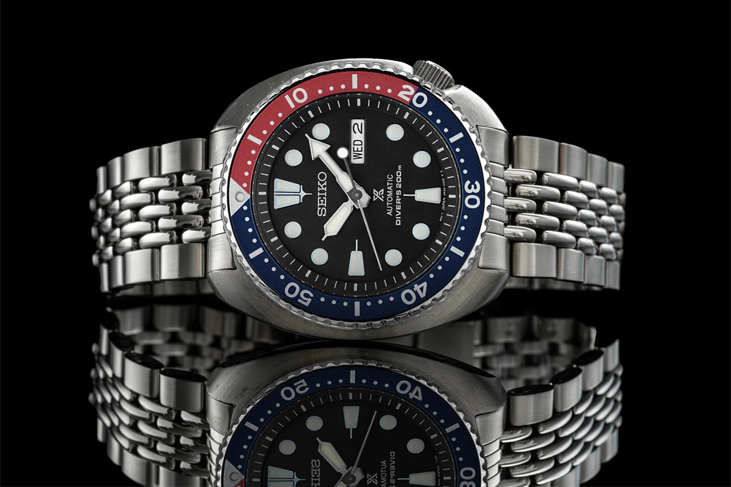 Seiko automatic dive watch with black dial, red and blue bezel, stainless steel bracelet, and date window against black background