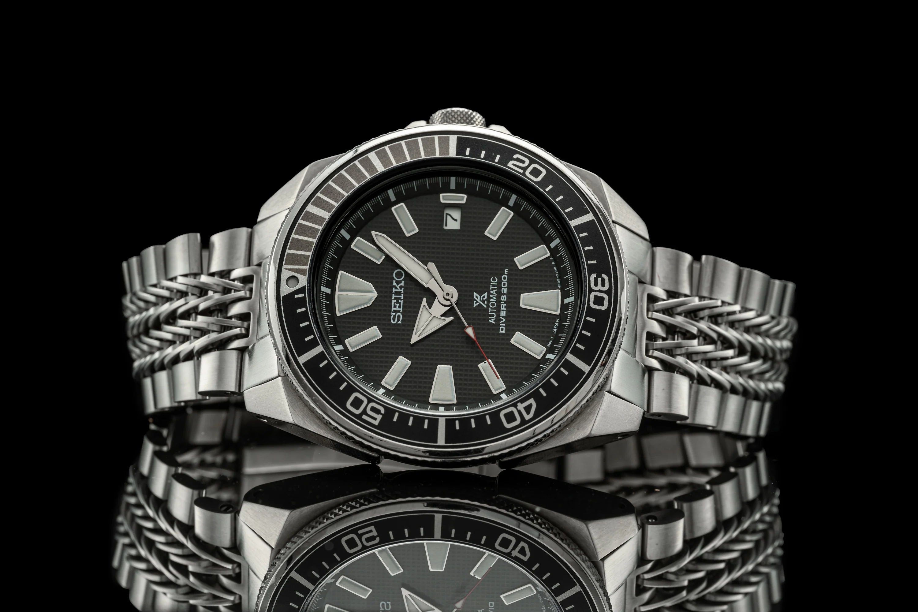 Seiko Samurai stainless steel dive watch with black dial, applied hour markers, and razor-wire metal bracelet on reflective black surface