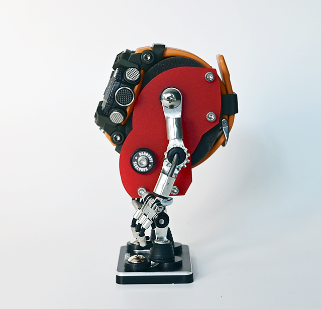 Red and black robot-shaped watch stand with articulated arms holding timepiece, featuring orange accents and chrome details on pedestal base