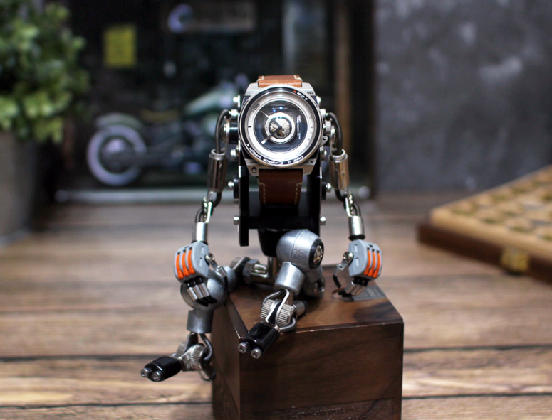 RoboMech silver metal watch stand with articulated robot arms holding brown leather strap watch on wooden base