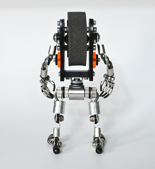 Mechanical robot watch stand with black textured platform, silver metal articulated arms and legs with orange accents, top view