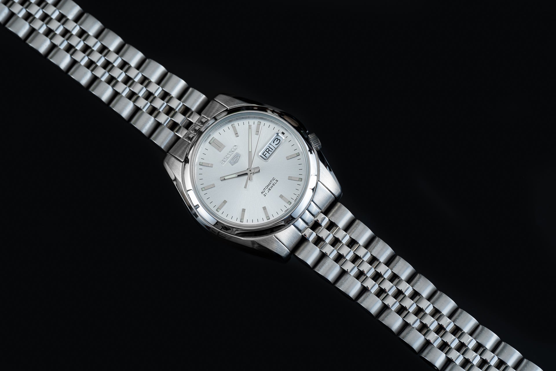Silver stainless steel automatic watch with white dial, day-date window, and matching metal bracelet on black background