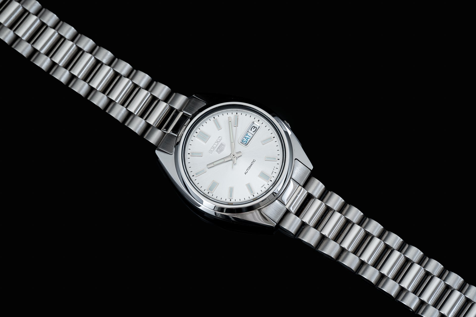 Silver stainless steel automatic watch with white dial, day-date display, and matching metal bracelet on black background