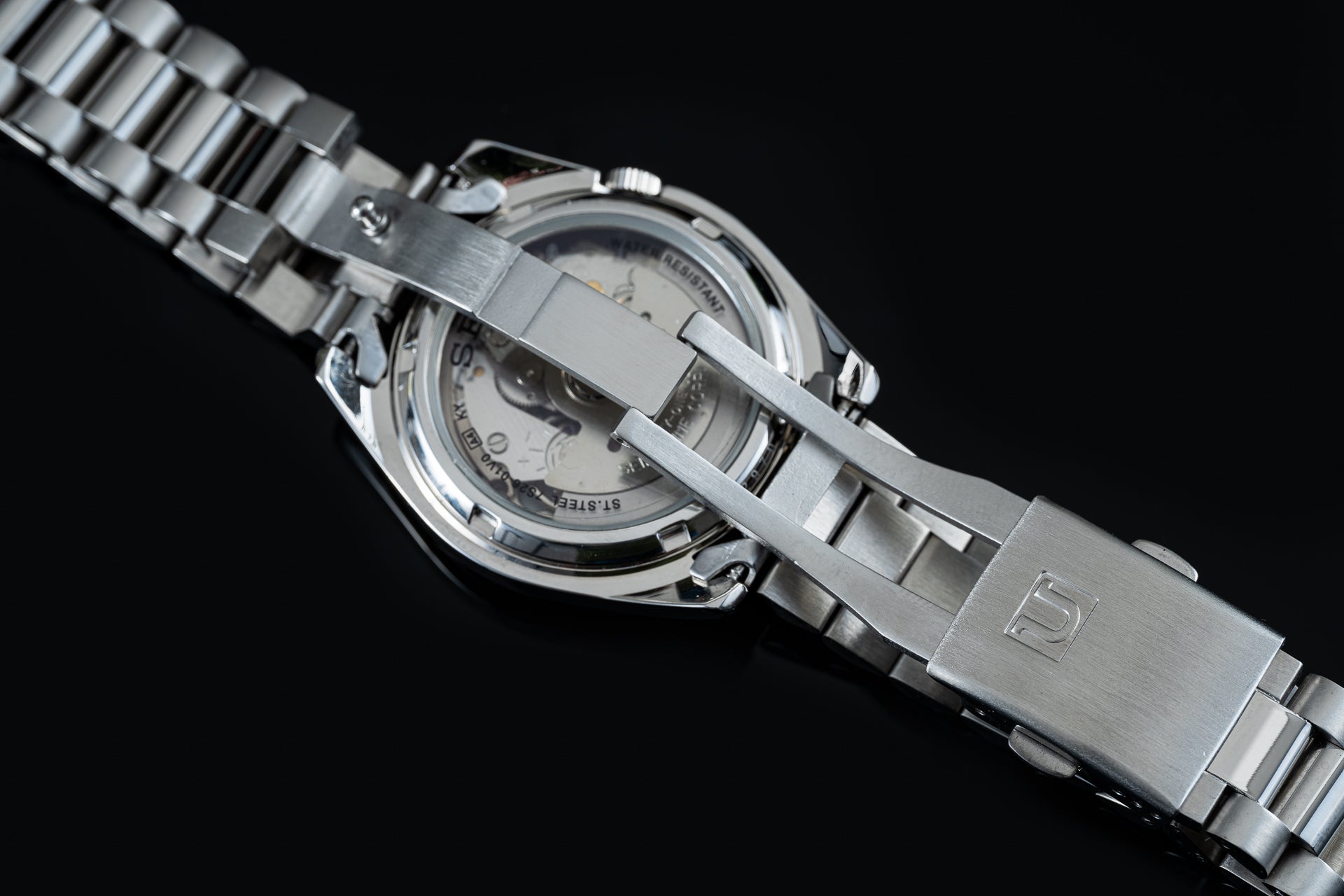 Silver stainless steel Seiko 5 dive watch with metal bracelet and open caseback revealing automatic movement on black background