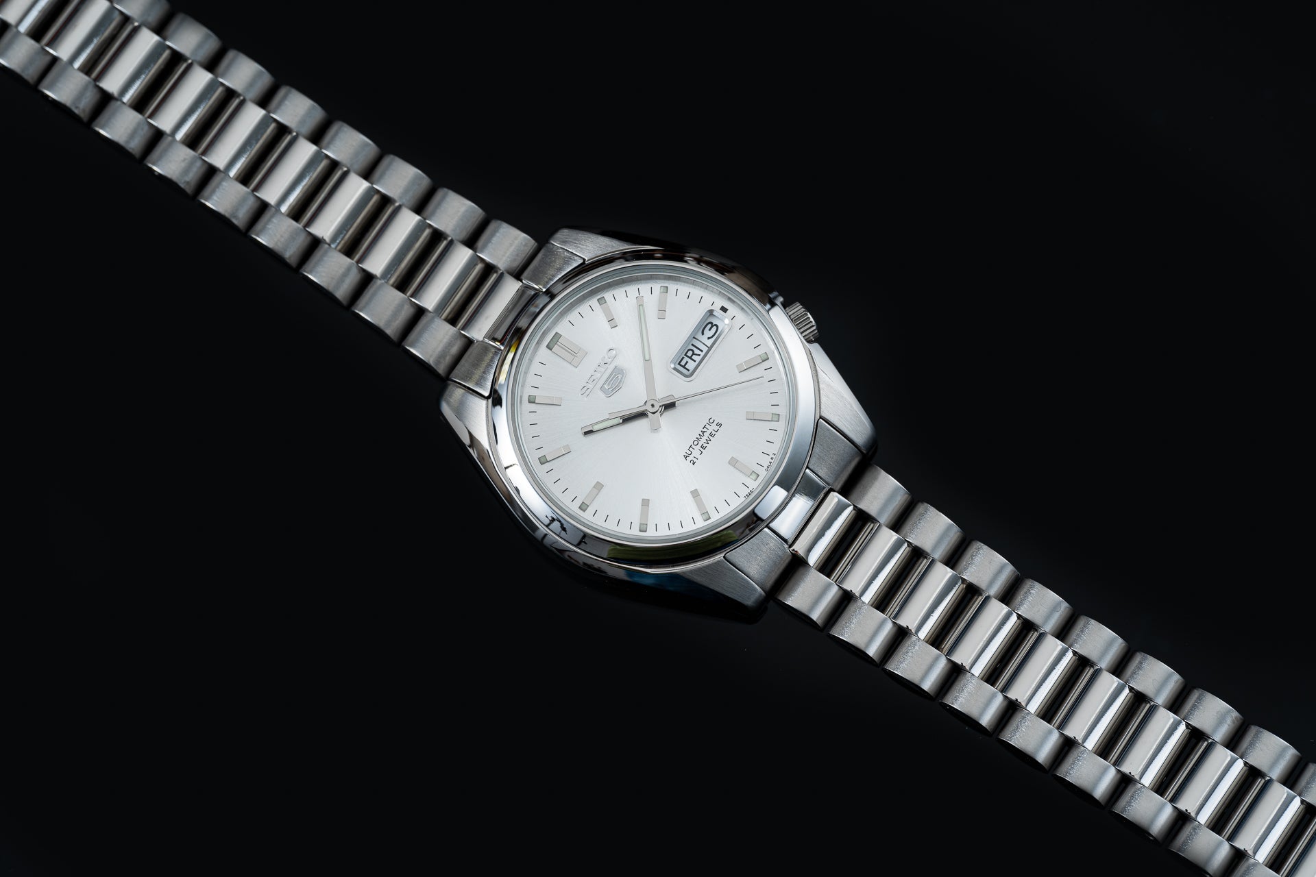 Silver stainless steel Seiko 5 automatic watch with white dial, day-date window, and matching metal bracelet on black background