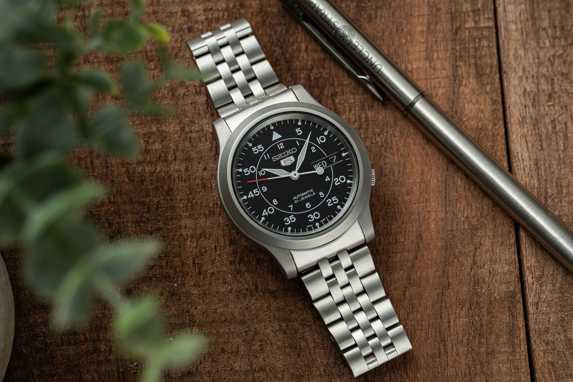 Seiko SNK809 automatic watch with stainless steel bracelet and black dial on wooden surface with pen
