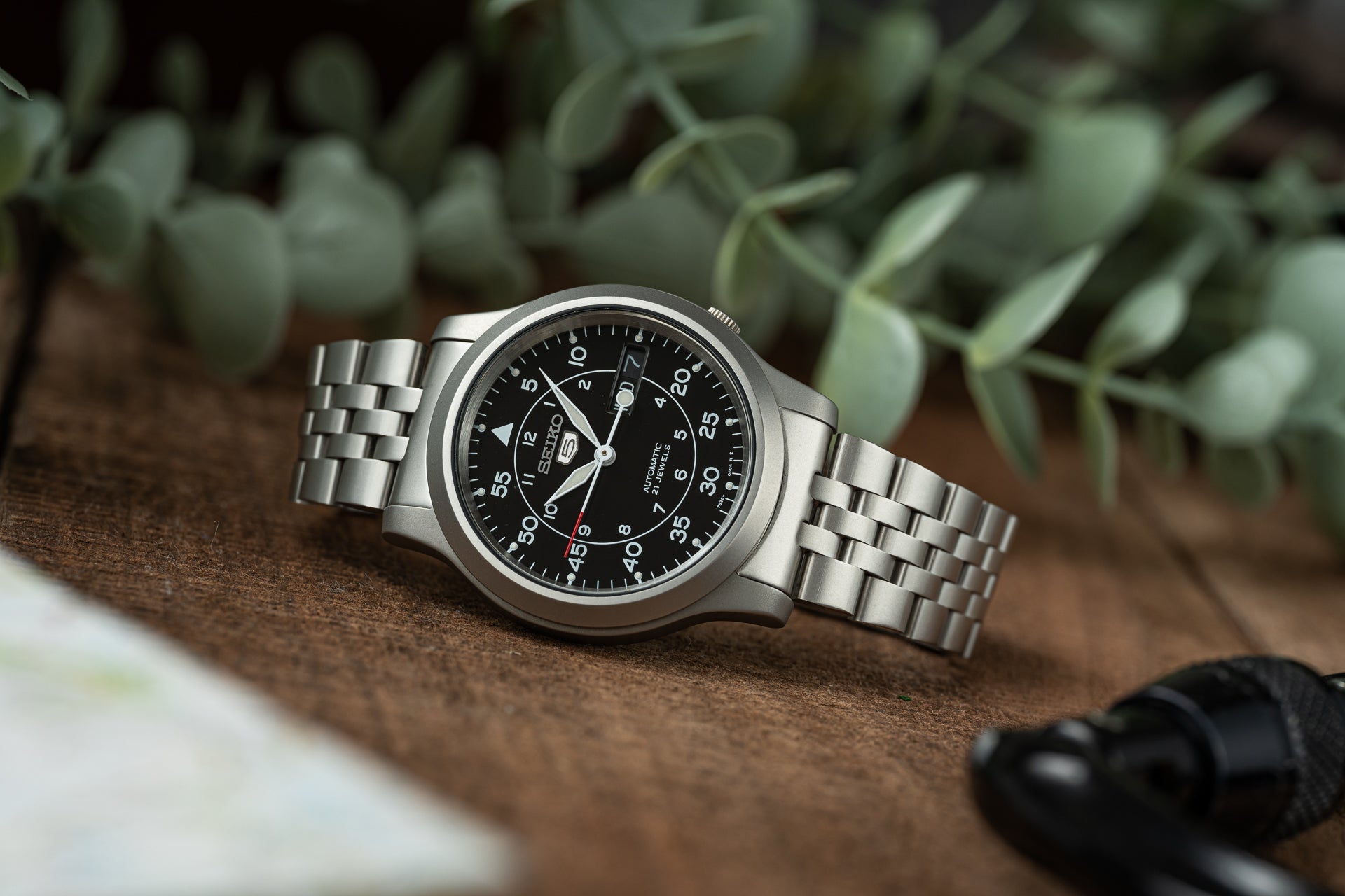 Seiko SNK809 automatic watch with stainless steel bracelet and black dial on wooden surface with green plant background