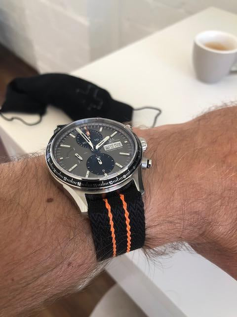 Stainless steel chronograph watch with black dial and day-date display on black orange stripe fabric strap, worn on wrist