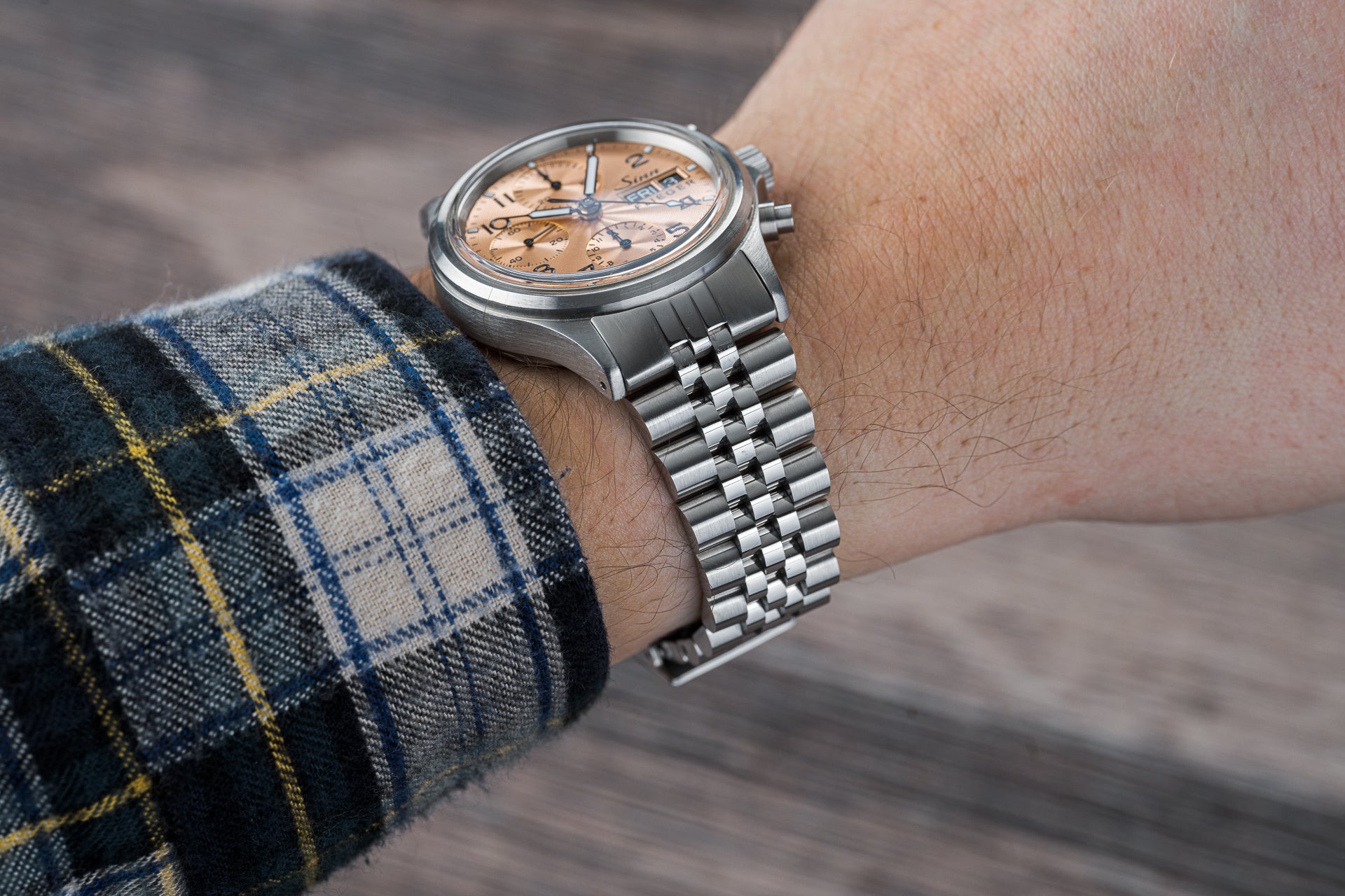 Stainless steel chronograph watch with salmon dial and metal bracelet worn on wrist with plaid sleeve