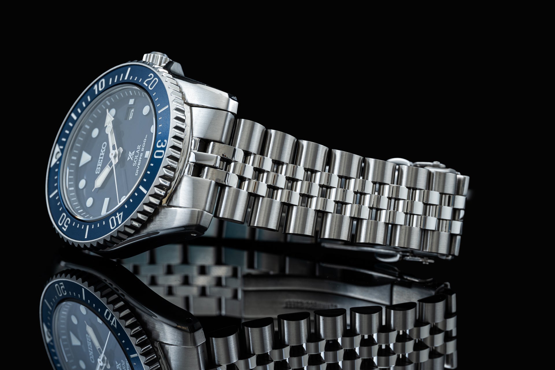 Seiko SNE57x solar dive watch with blue dial, rotating bezel, and stainless steel bracelet on black background