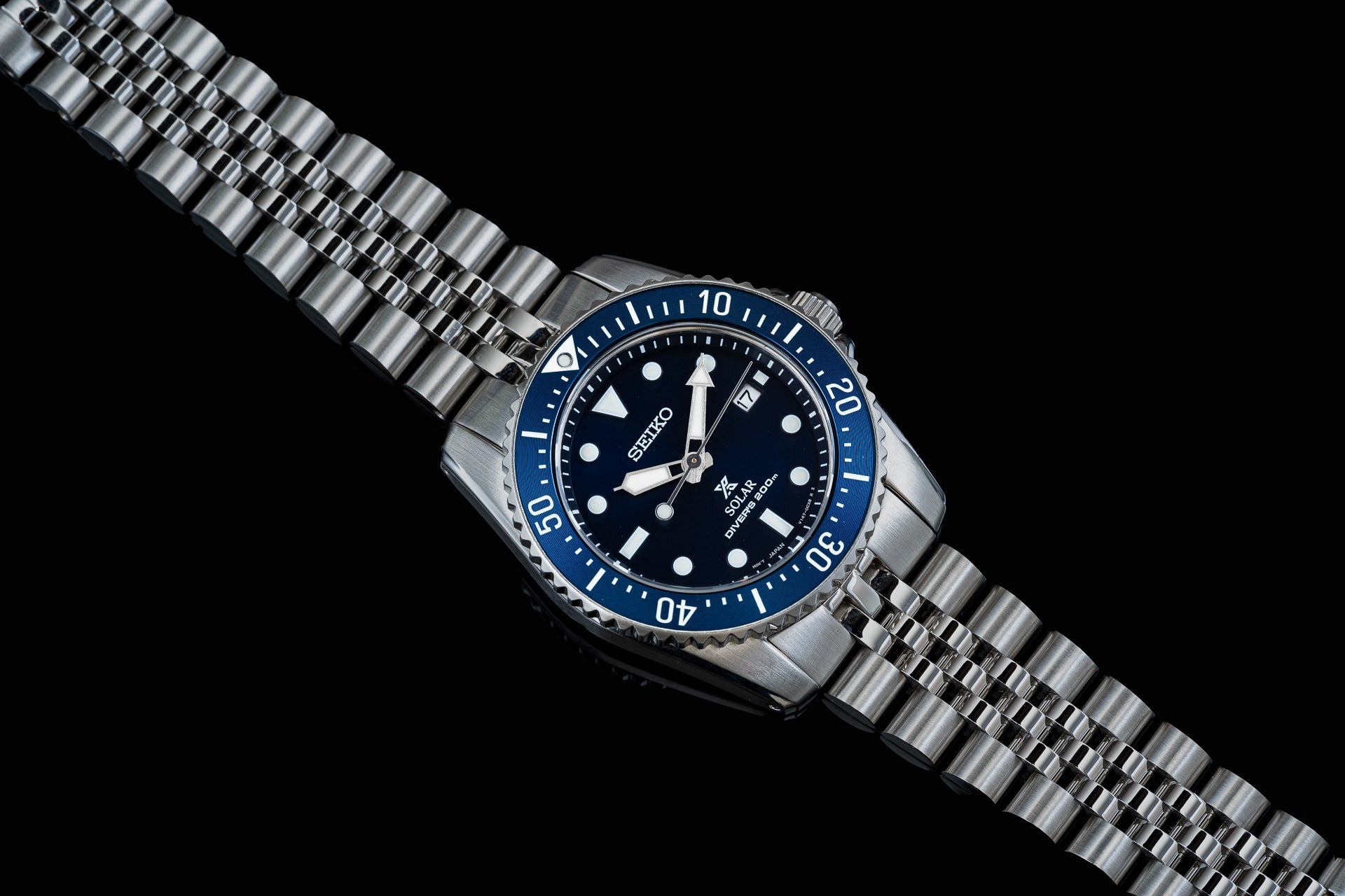 Seiko SNE57x solar dive watch with blue bezel, black dial, stainless steel bracelet and date window on black background