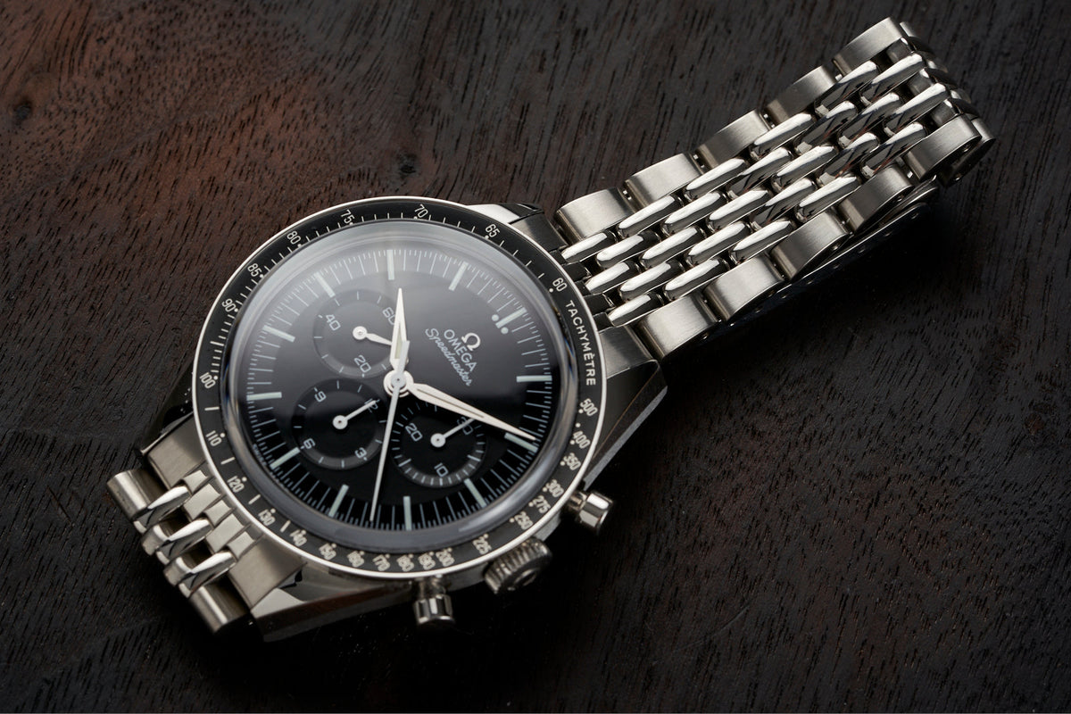 Omega Speedmaster chronograph watch with black dial, tachymeter bezel, and stainless steel Beads of Rice bracelet on dark wood surface