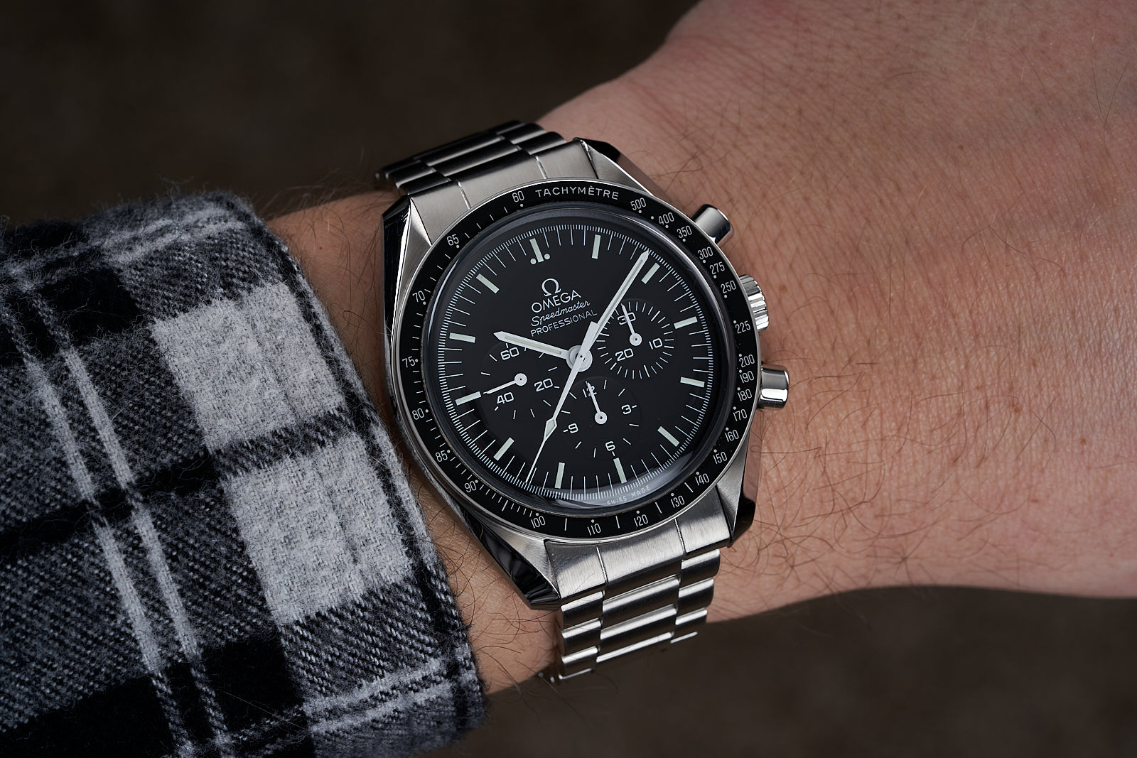 Omega Speedmaster Professional chronograph watch with black dial and stainless steel bracelet worn on wrist with plaid sleeve