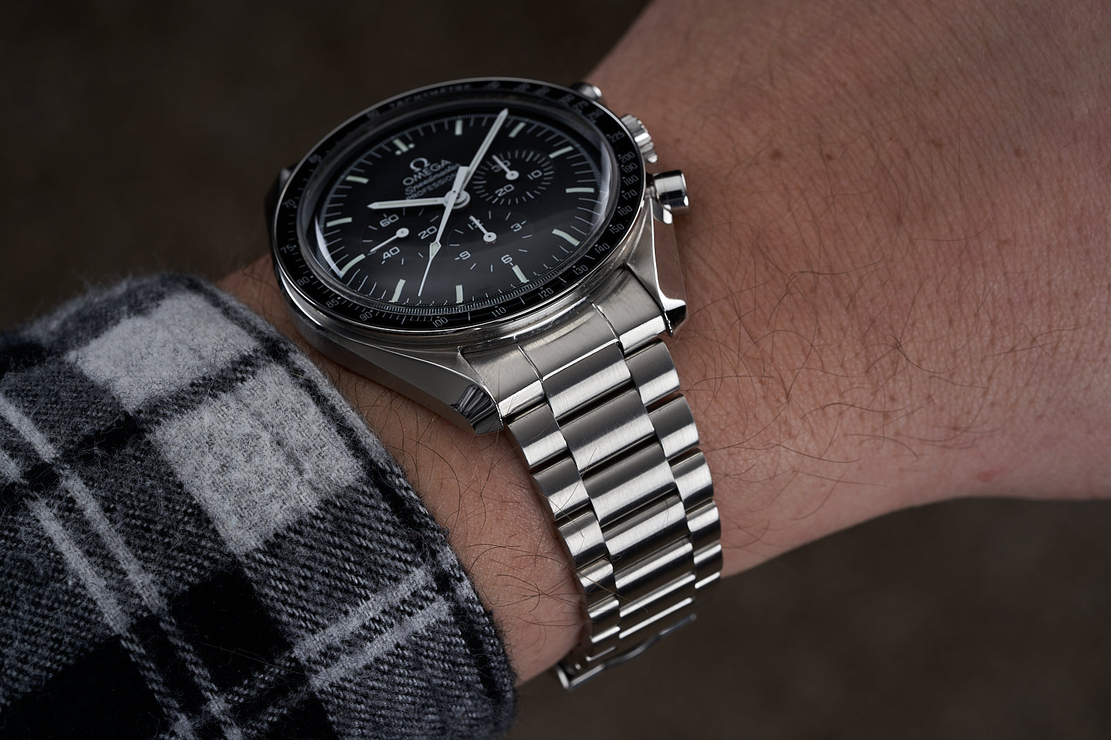 Omega Speedmaster chronograph watch with black dial and stainless steel bracelet worn on wrist with plaid shirt sleeve