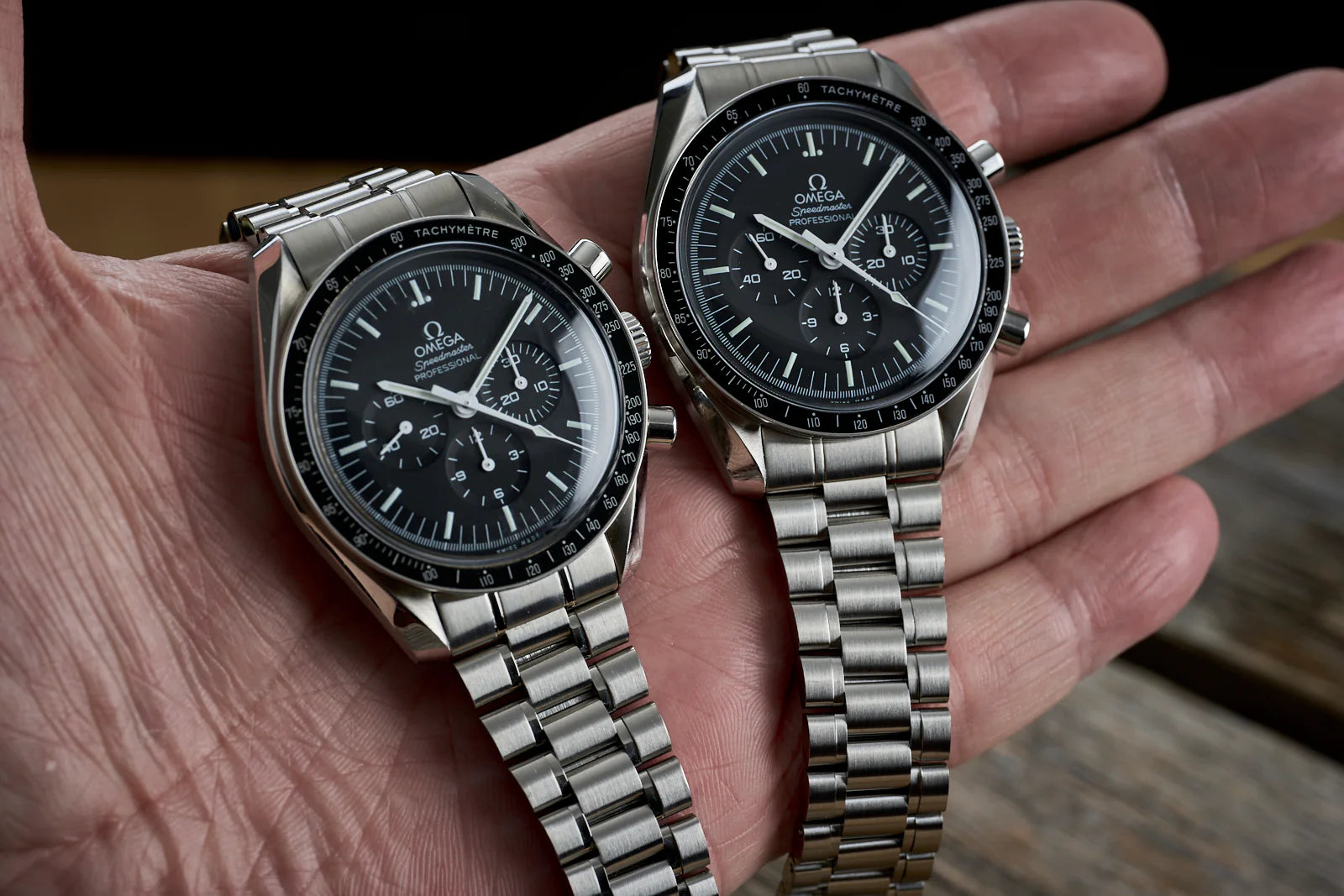 Two Omega Speedmaster chronograph watches with stainless steel bracelets and black dials held in hand, showing tachymetre bezel and subdials