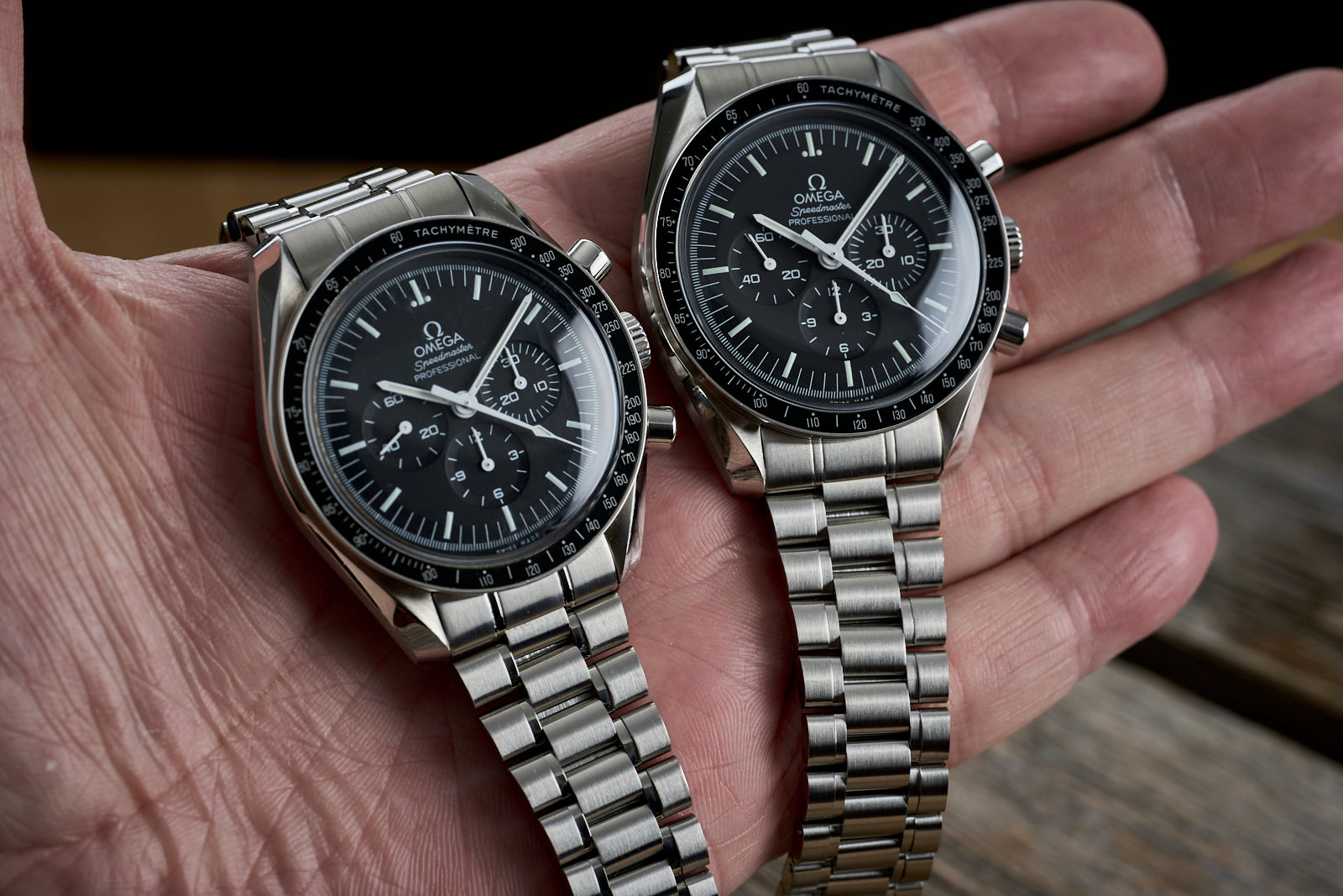 Two Omega Speedmaster chronograph watches with black dials and tachymeter bezels on stainless steel bracelets held in hand