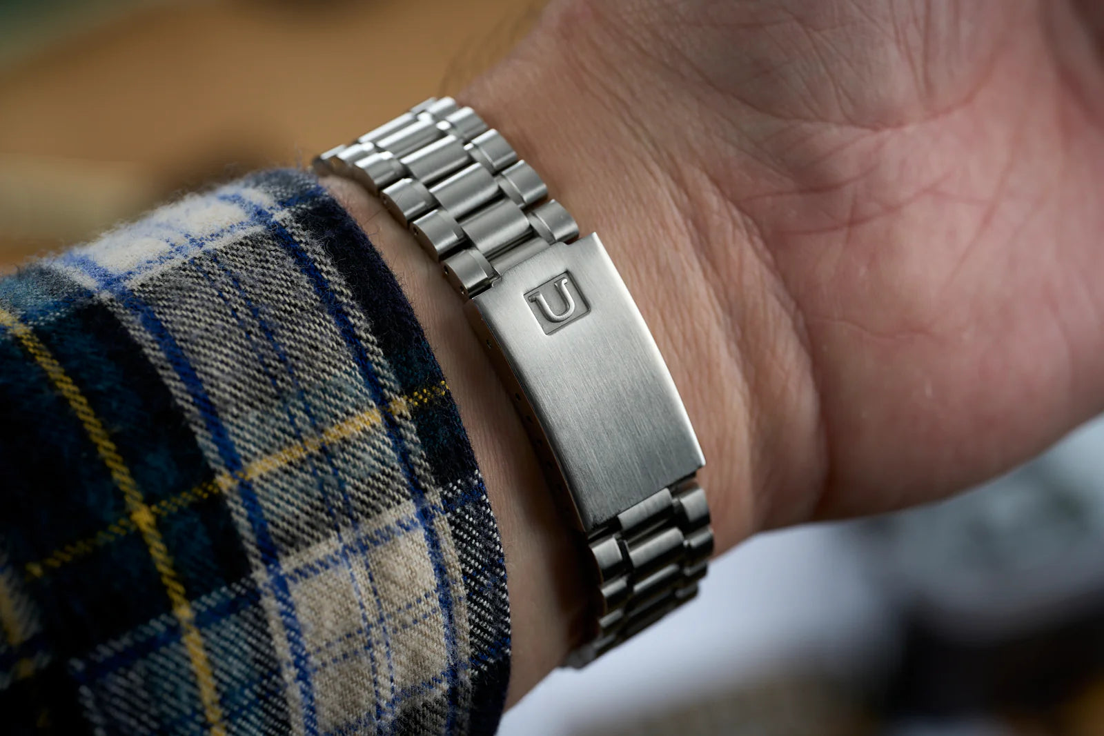 Stainless steel link bracelet with brushed finish and engraved U logo, worn on wrist with blue plaid shirt