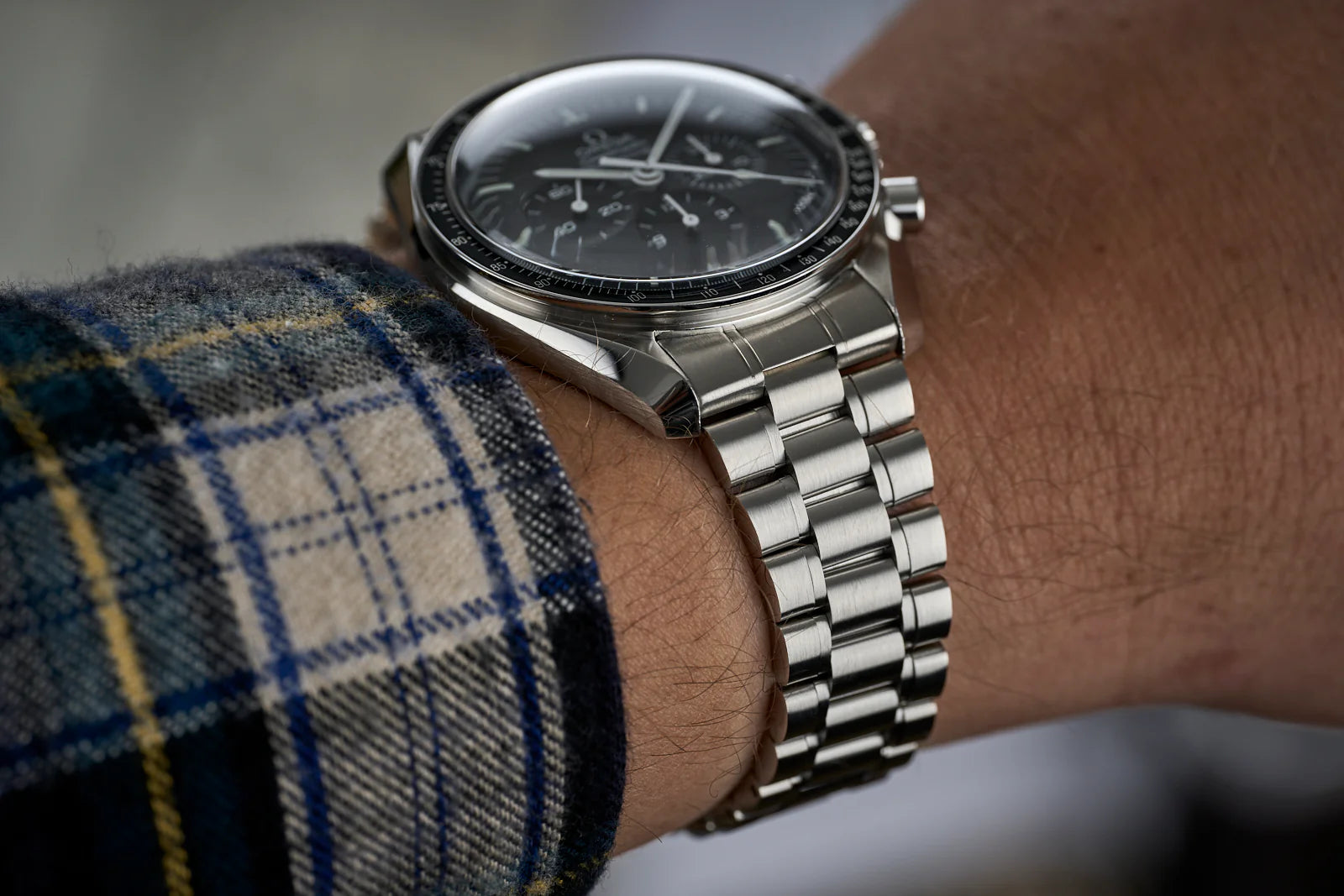 Omega Speedmaster chronograph watch with black dial and stainless steel bracelet worn on wrist with plaid shirt sleeve