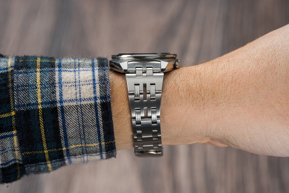 Stainless steel sport watch bracelet on wrist with plaid shirt sleeve, showing polished metal links and secure clasp fastening